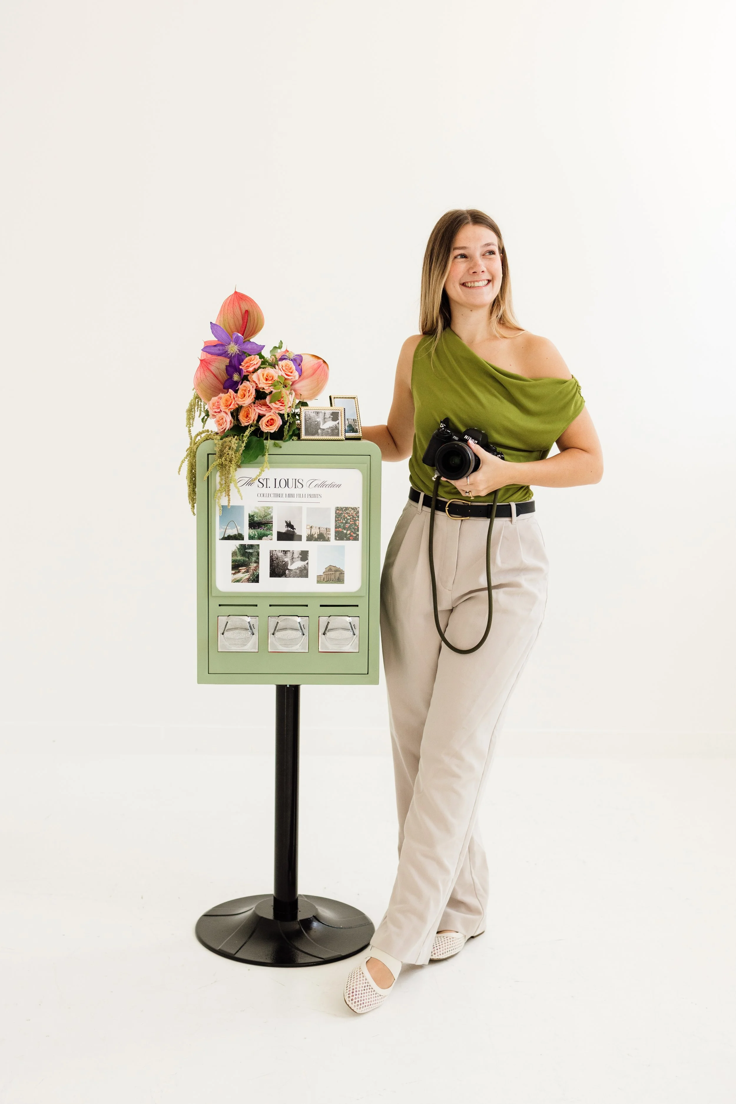 A woman standing next to a display stand with flowers and photographs, holding a camera in her right hand and smiling.