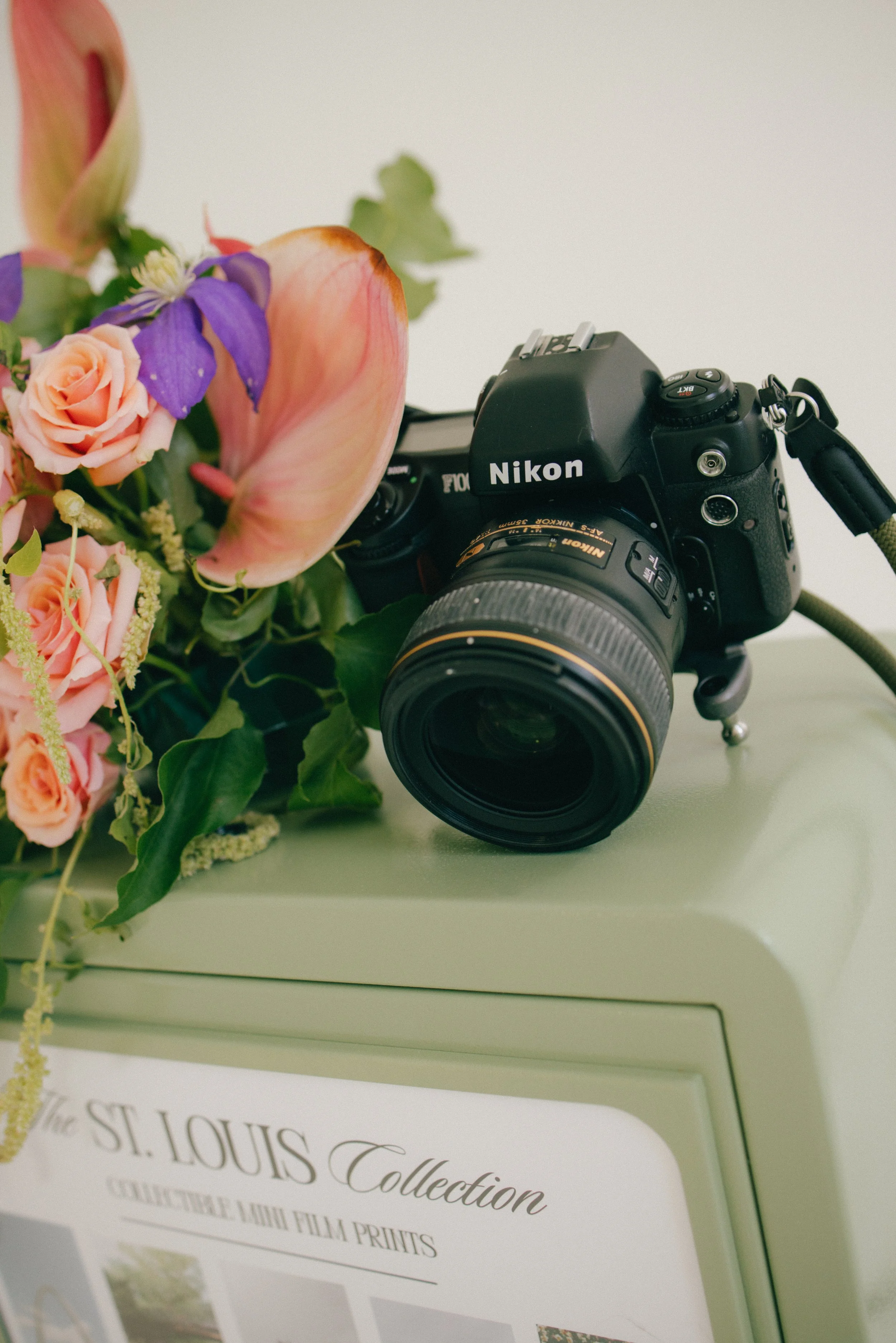 A close-up of a black Nikon camera placed on a light green surface next to a bouquet of pink roses, purple flowers, and green foliage, with a white background and a partly visible printed paper in the foreground.