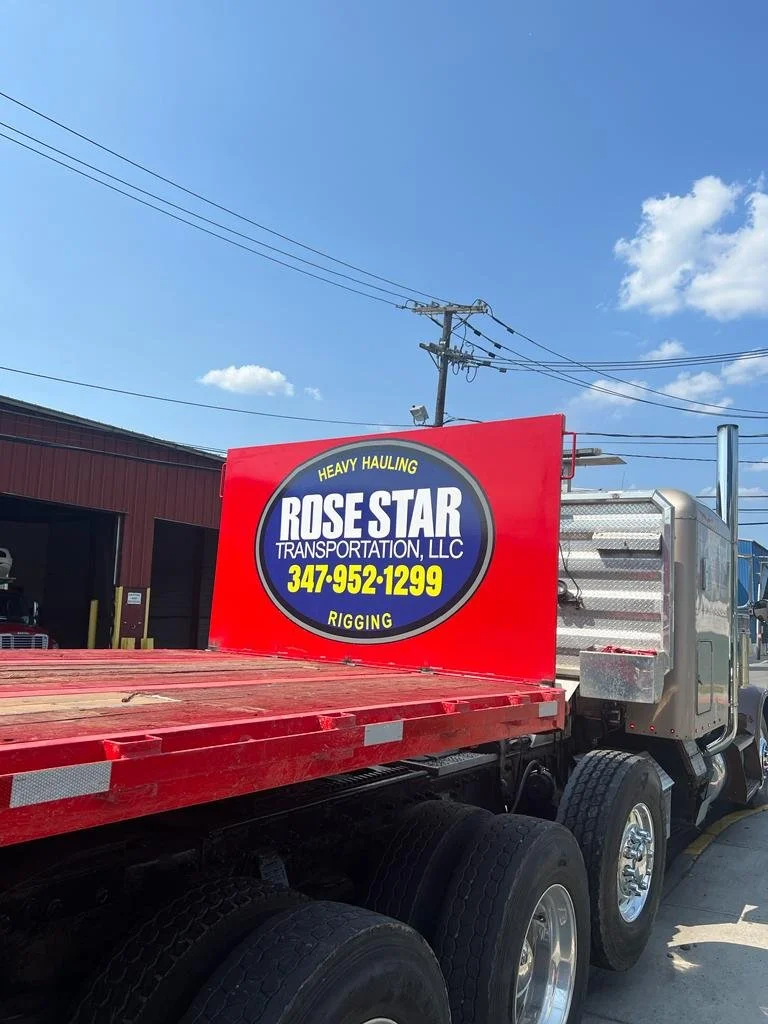 Red flatbed truck with a sign reading Rose Star Transportation LLC, heavy hauling, rigging, phone number 347-952-1299, parked in a lot with a blue sky and some clouds overhead.