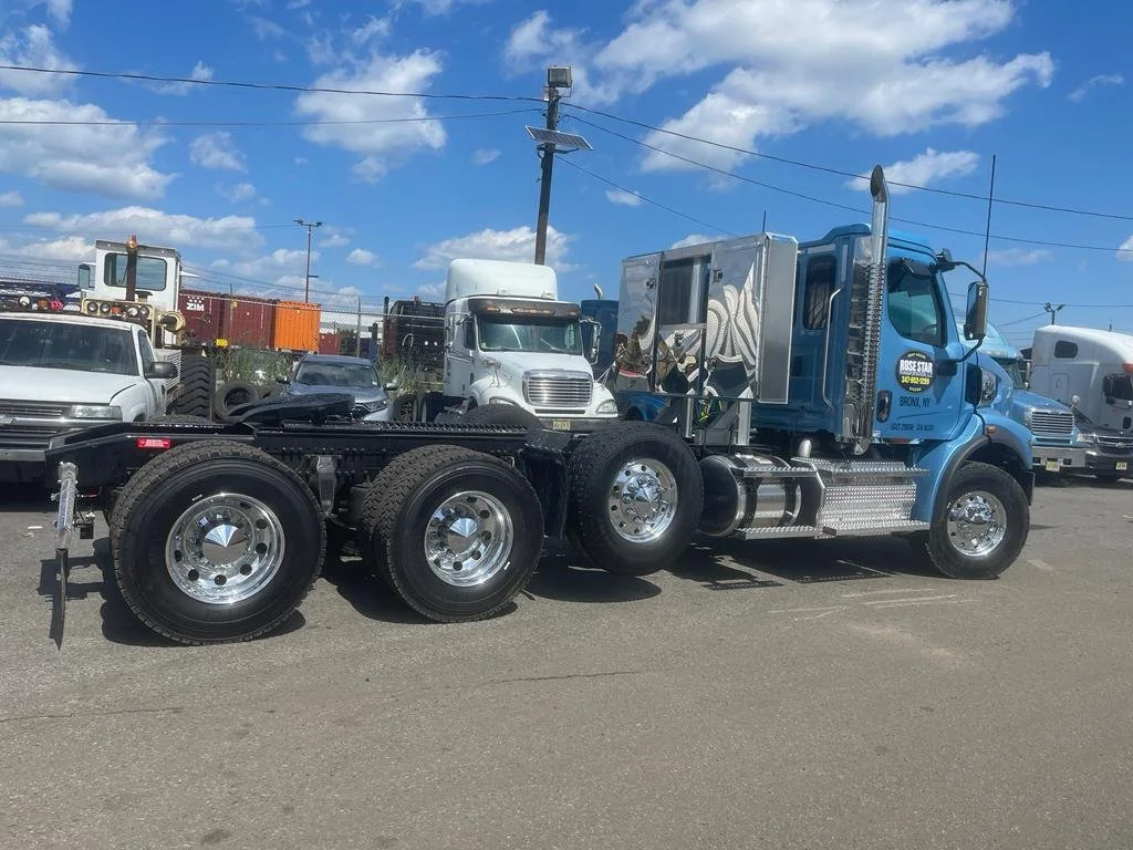Blue semi-truck with no trailer attached parked in a lot with other trucks and cars under a partly cloudy sky.