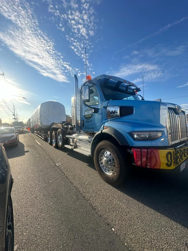 Blue semi-truck carrying a load on a busy road with other cars, under a partly cloudy sky during daylight.