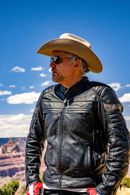 A man wearing a cowboy hat, sunglasses, and a leather jacket stands outdoors against a clear blue sky with the Grand Canyon in the background.