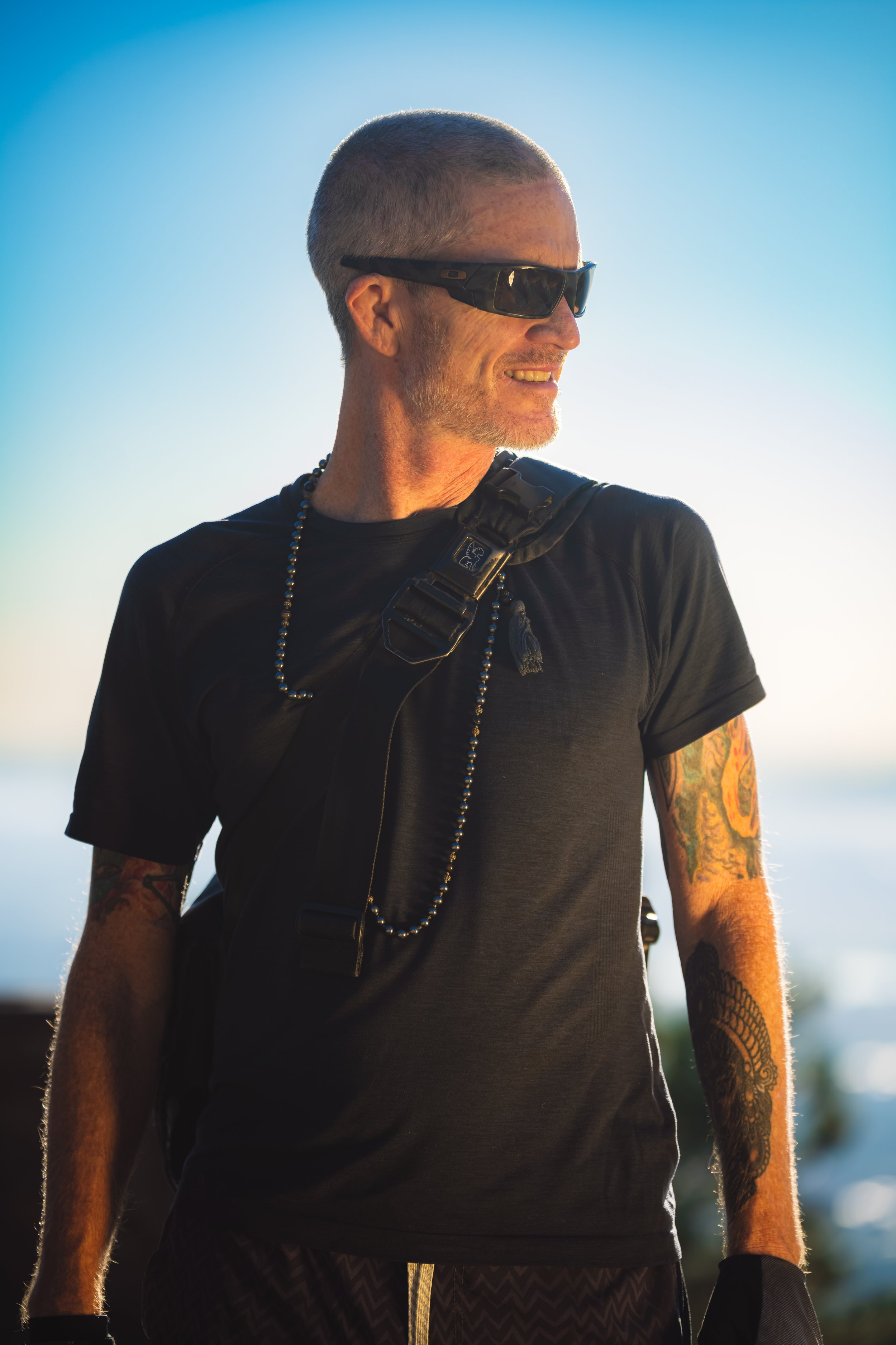 A man with short gray hair and a beard, wearing black sunglasses, a black t-shirt, and tattoos on his arms, smiling outdoors during daylight with a clear sky background.
