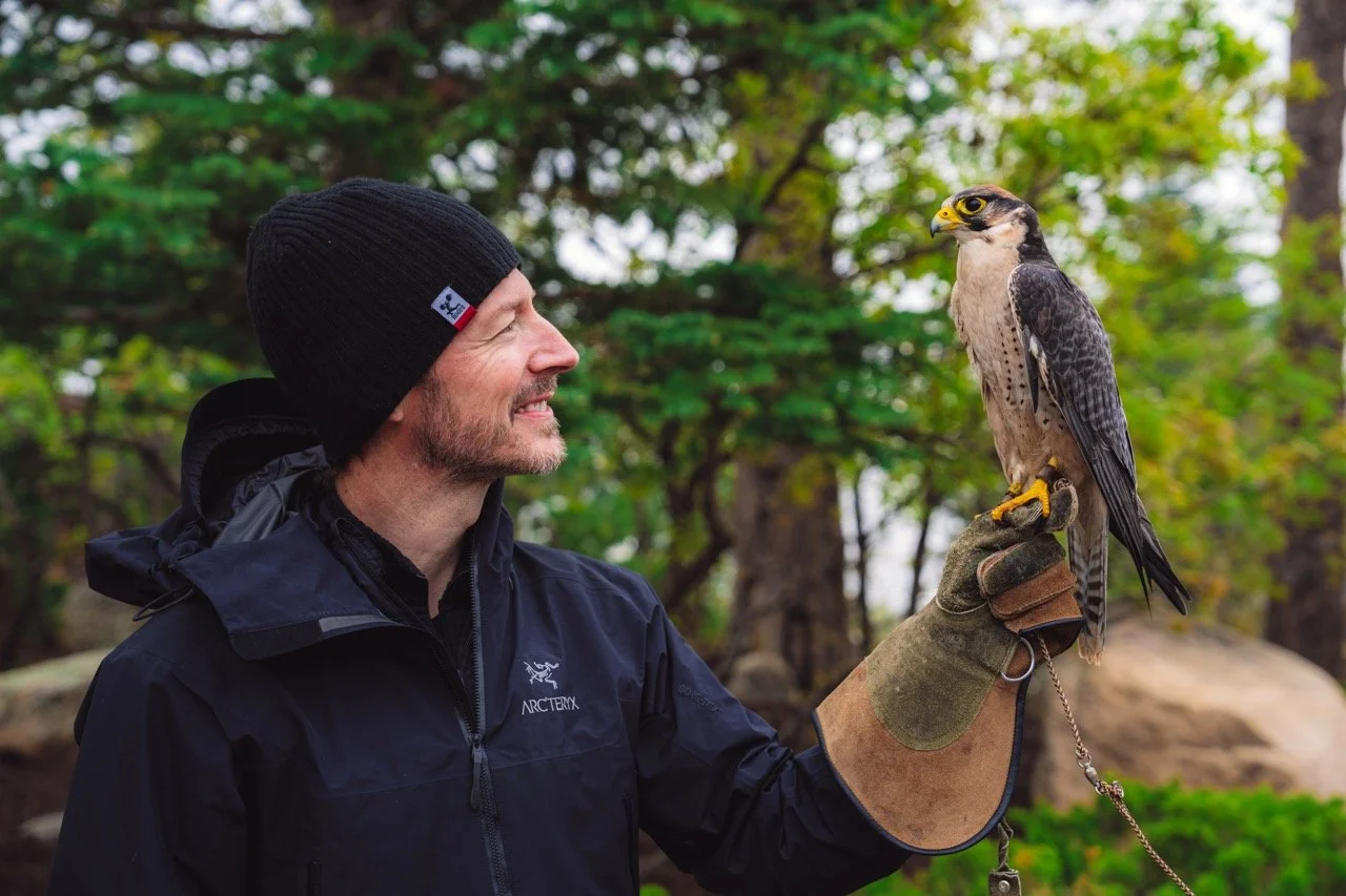 A man in outdoor gear smiling at a falcon perched on his gloved hand in a wooded area.