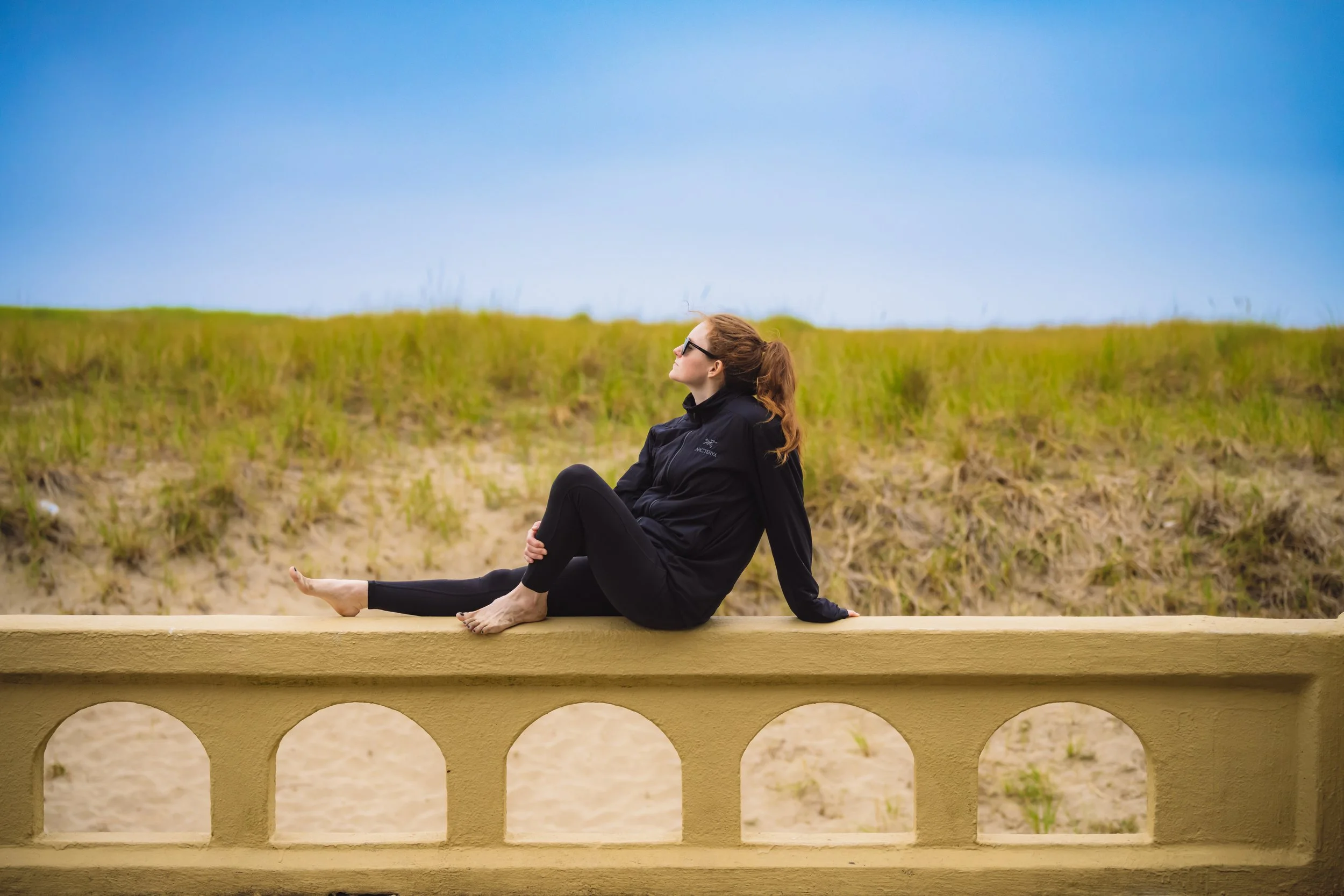 A woman in black athletic clothing sitting on a yellow wall, with a grassy hill and blue sky in the background.