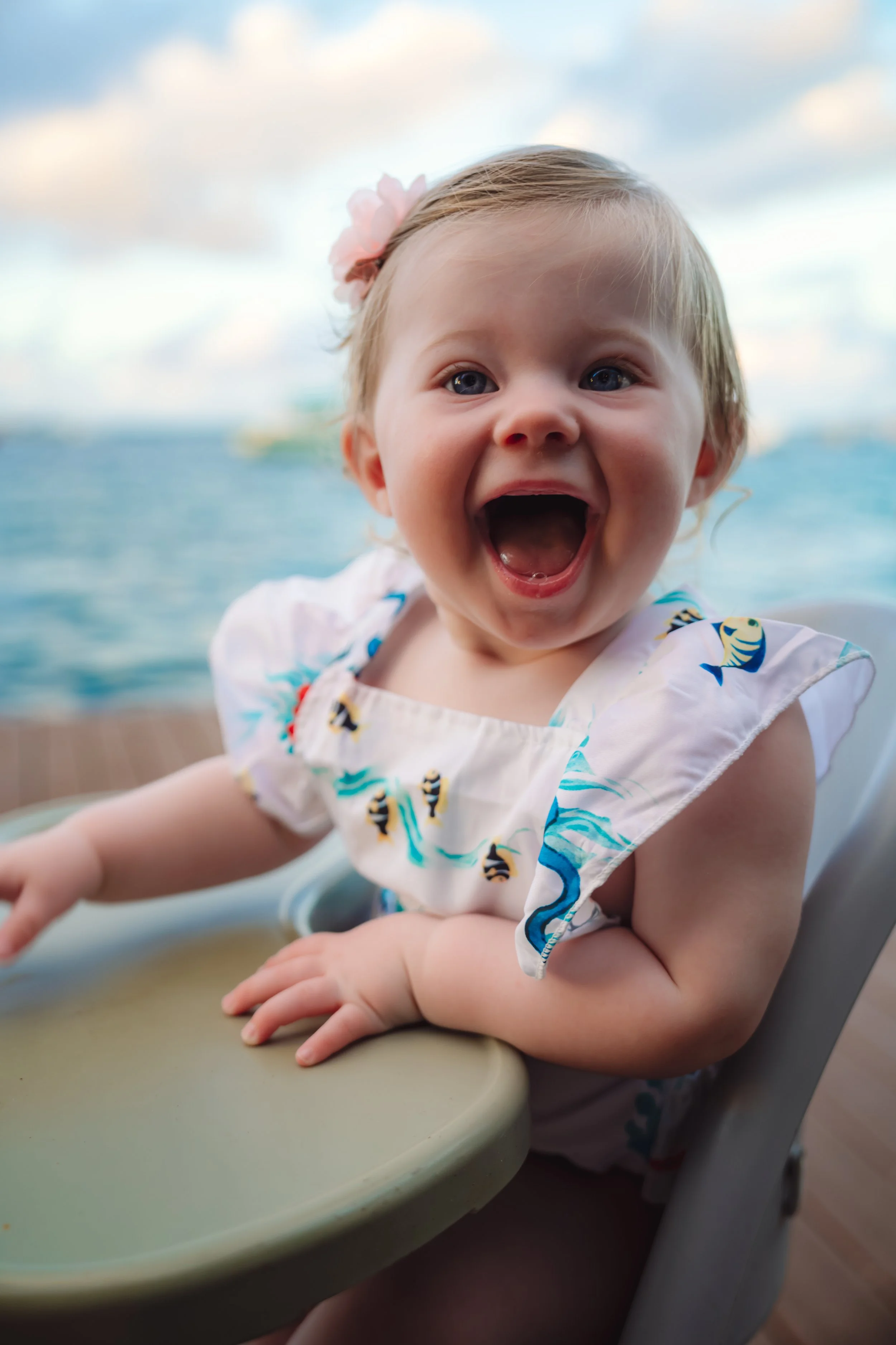 A happy toddler girl with a pink flower hair accessory, sitting in a high chair outdoors by the water, smiling widely with her mouth open.