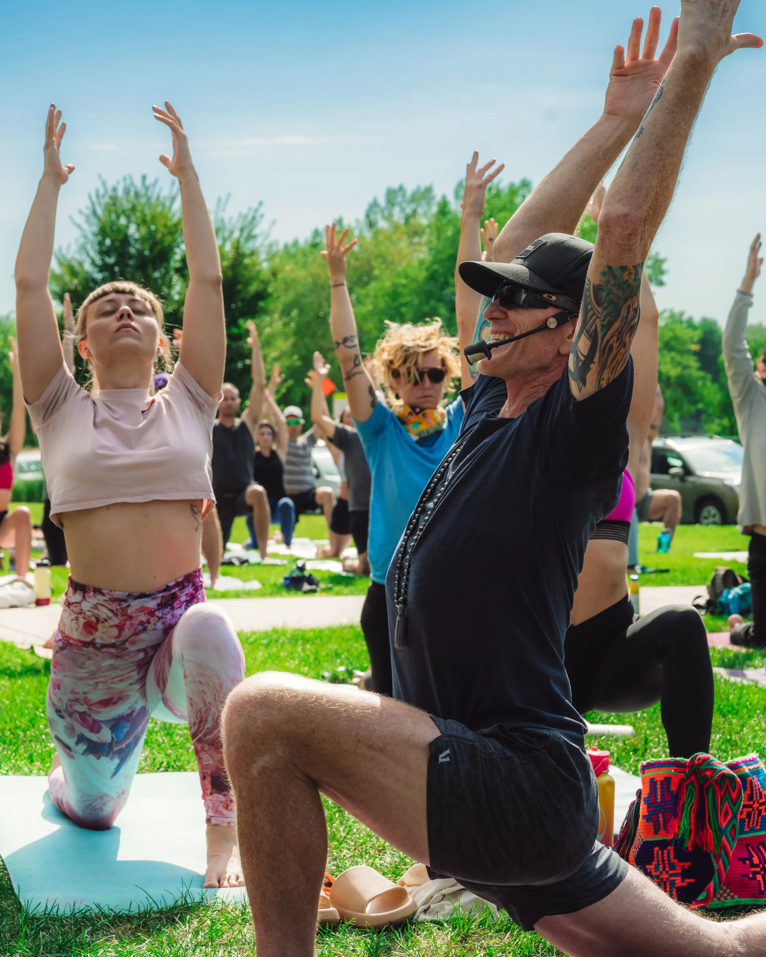 Rob Loud wearing a headset and sunglasses, leading a group engaged in yoga outdoors in a park, raising their arms in a yoga pose. 