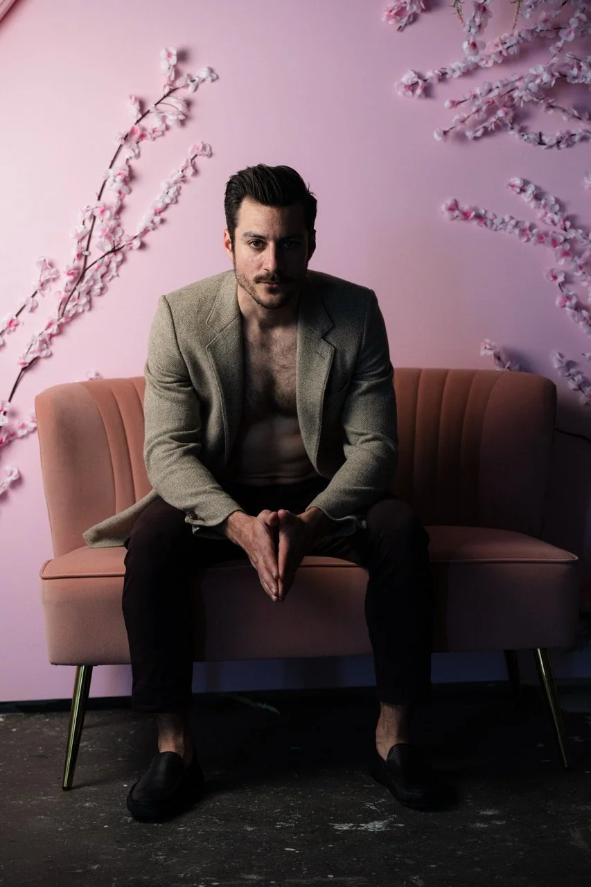 A man sitting on a pink sofa in front of a pink wall decorated with cherry blossom branches, dressed in a gray blazer, black pants, and black shoes, with a serious expression.