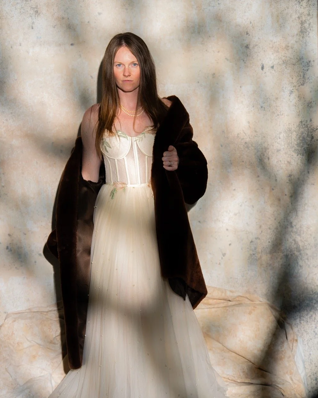 A woman with long, straight brown hair wearing a white vintage style dress and a string of pearls, draped in a dark fur coat over her shoulders, standing against a textured beige background.