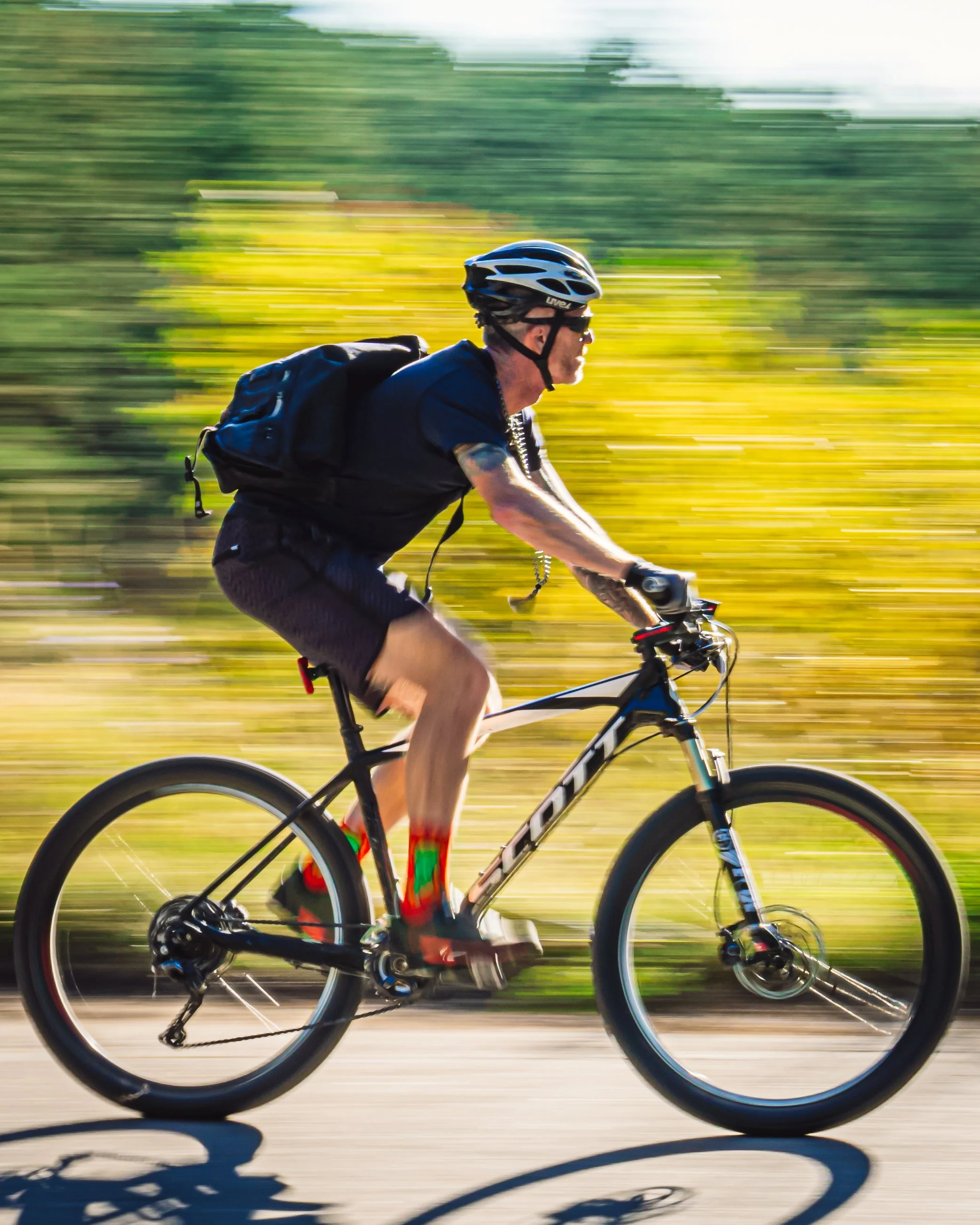 Rob Loud riding a mountain bike on a dirt trail during daytime, wearing a helmet, sunglasses, black t-shirt, and shorts, with a backpack, in a blurred outdoor setting.