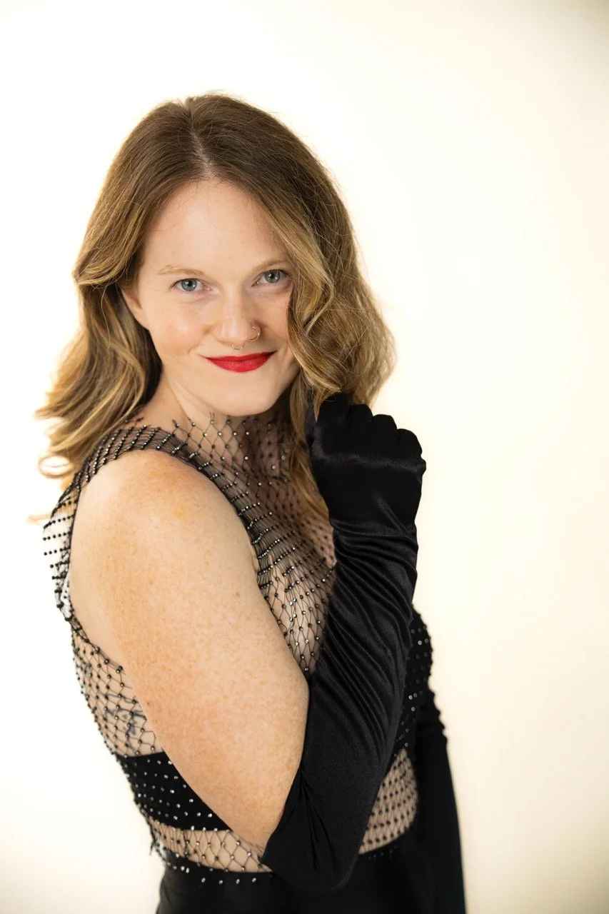 A woman with shoulder-length red hair, blue eyes, and fair skin, wearing a black fishnet top with embellishments, black gloves, and red lipstick, looking at the camera with a slight smile.