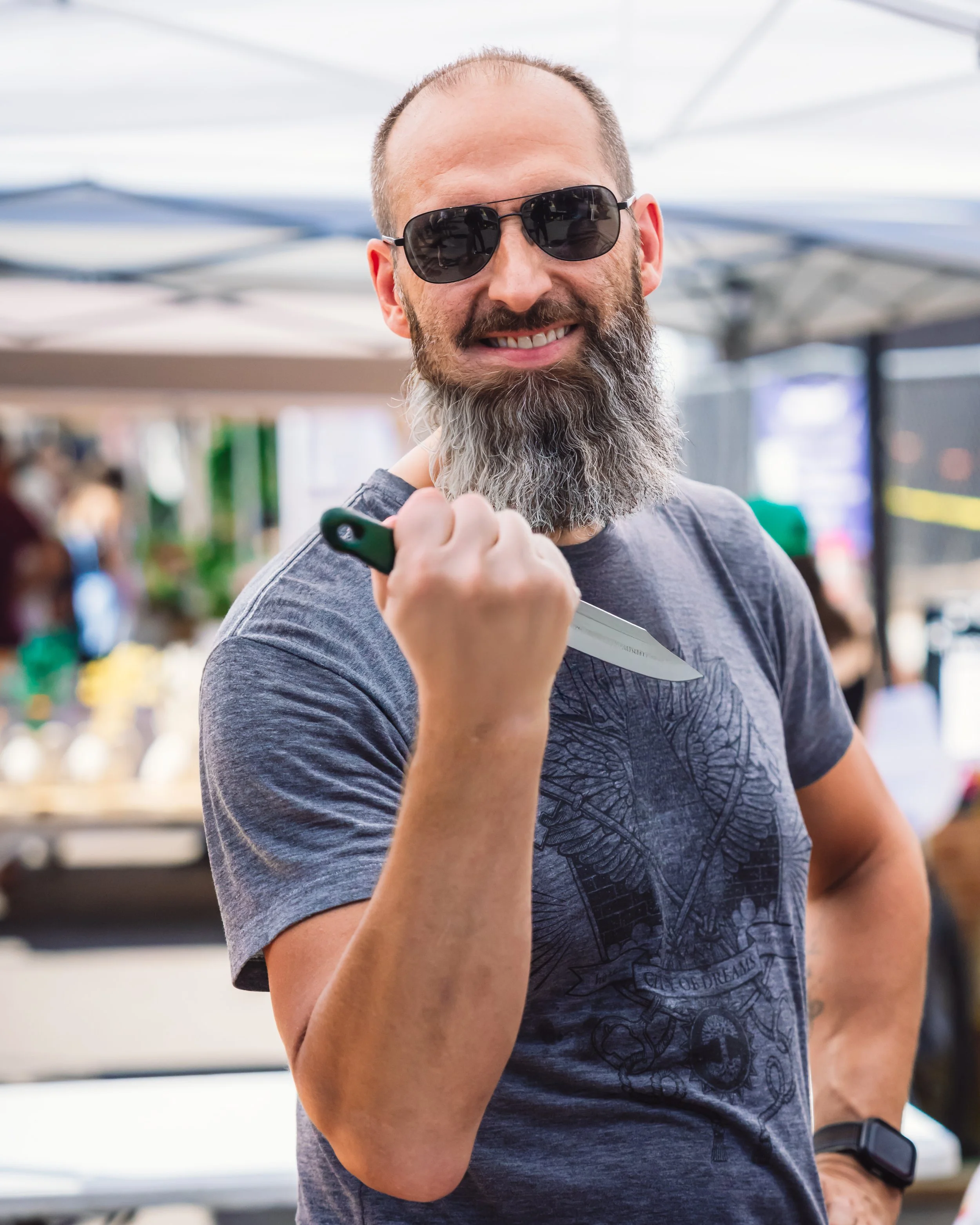 A bearded man wearing sunglasses and a gray t-shirt is holding a kitchen knife with a confident smile as he prepares to sharpen it. The background shows an outdoor market.