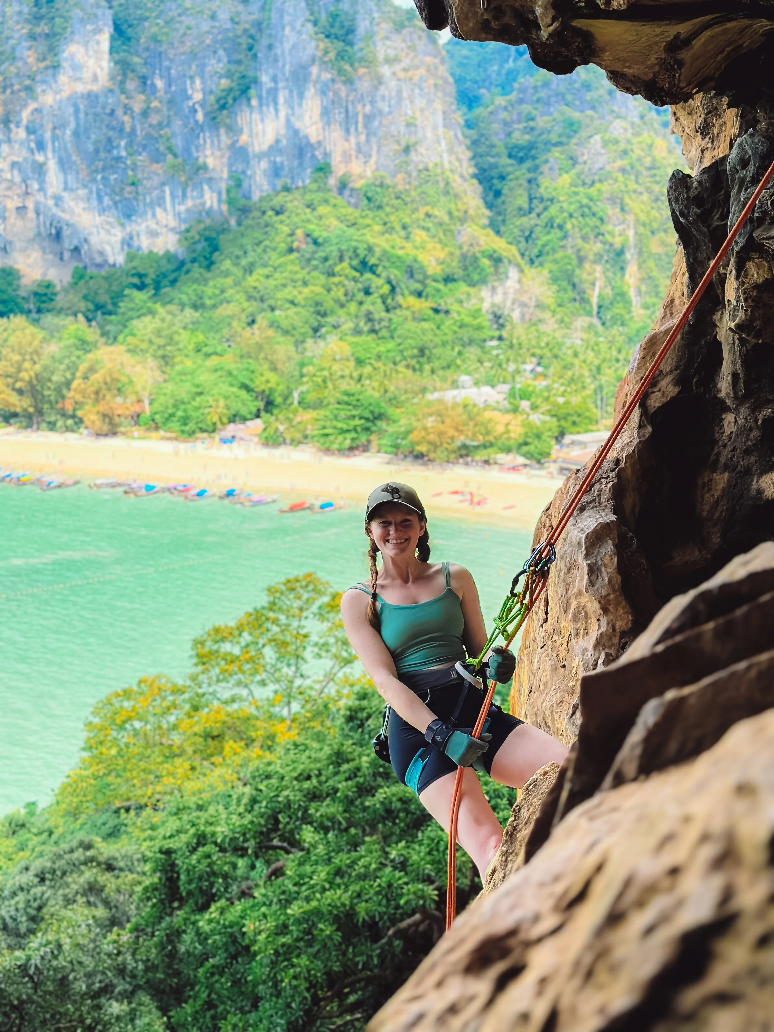 A woman rock climbing on a cliffside in Railay Beach, Thailand with a scenic background of a beach, trees, and mountains, wearing a helmet, harness, and outdoor clothing.
