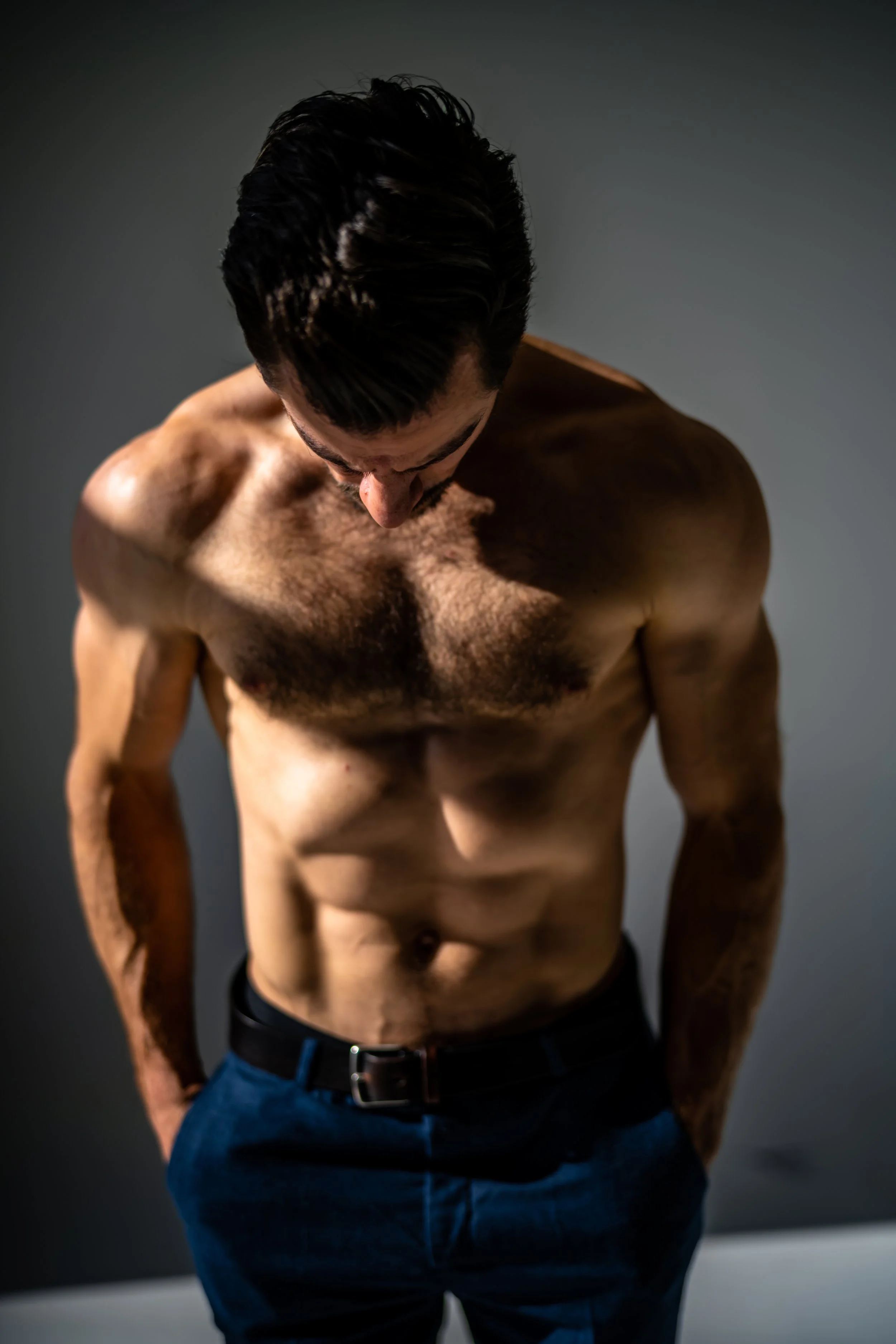 A shirtless man with dark hair is looking down, against a dark gray background.
