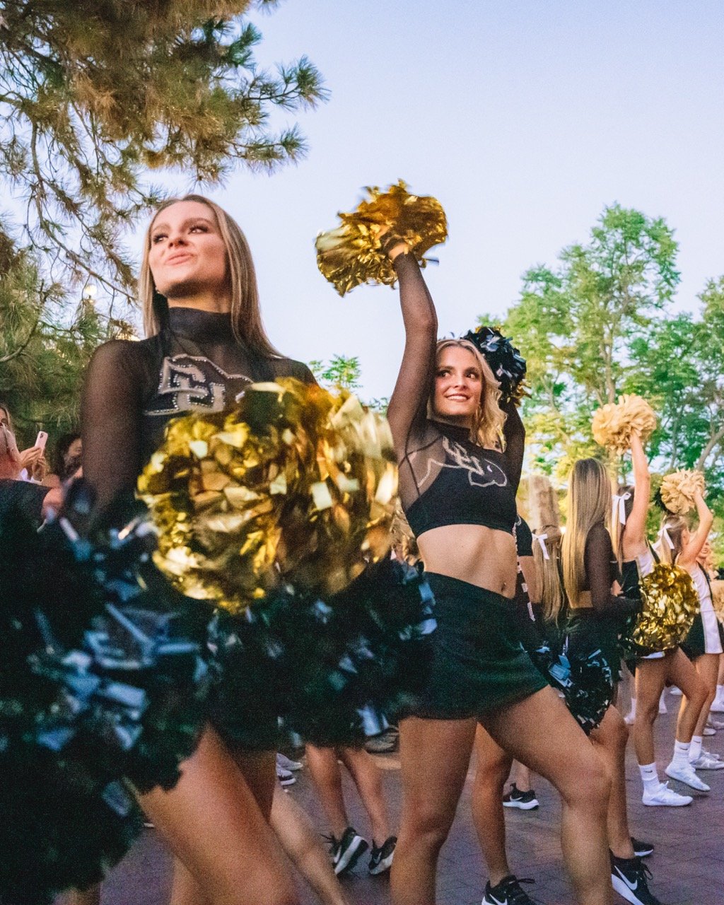 University of Colorado cheerleaders performing outdoors with pom-poms, wearing black outfits with tree and sky background.