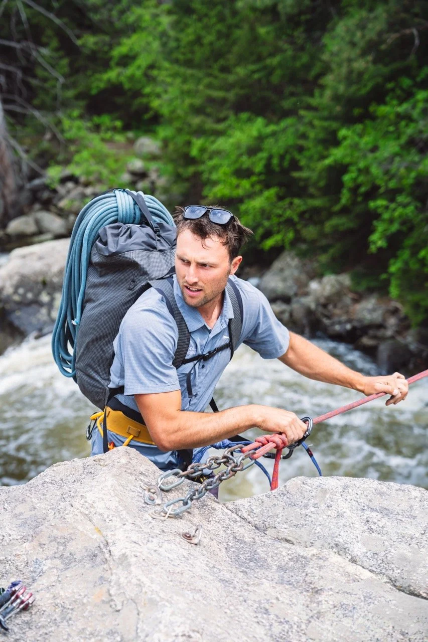 A dark-haired man dismounting from a Tyrolean traverse after crossing Boulder Creek, CO, carrying a backpack with a coiled rope, with a concerned expression.