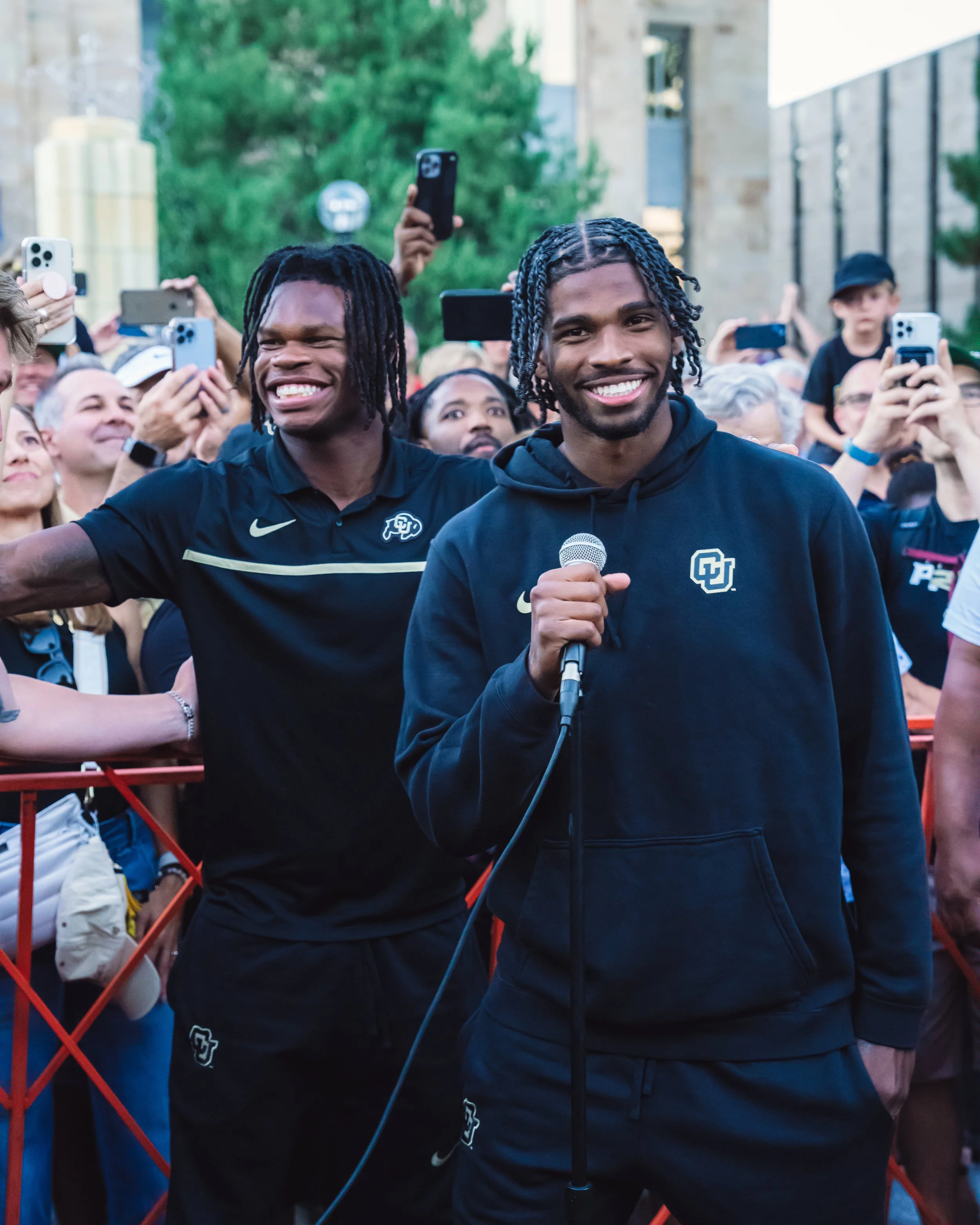 Two young men, one holding a microphone, smiling at a crowd during an outdoor event, with many people taking photos.