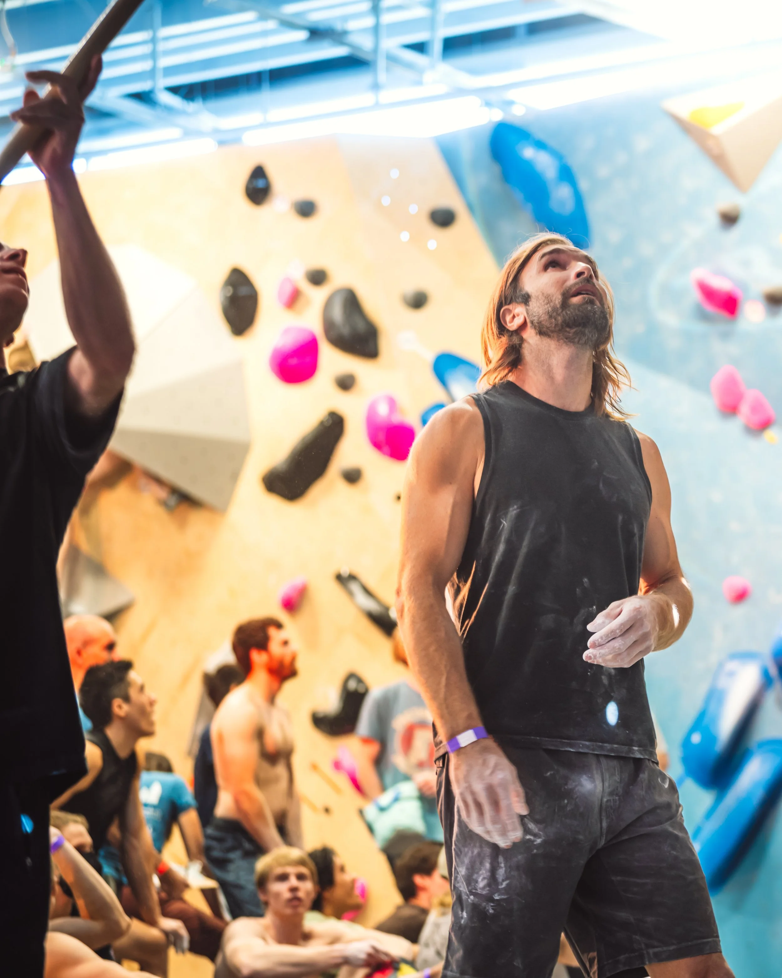 Chris Sharma looking up at the climbing wall for a competition at G1 Climbing and Fitness, surrounded by other climbers.