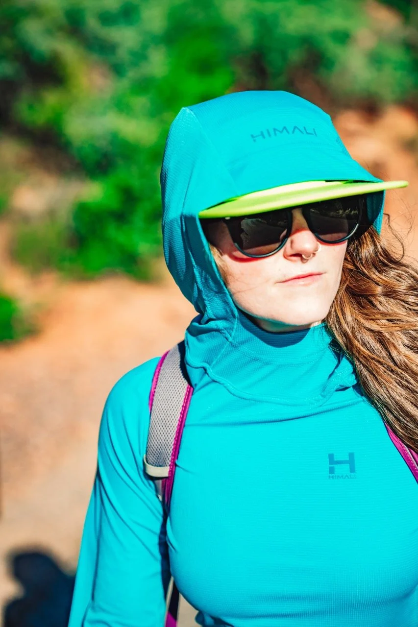 A woman in outdoor hiking gear wearing a blue hooded jacket, a blue sun hat with a green brim, sunglasses, and a backpack on a trail surrounded by greenery.