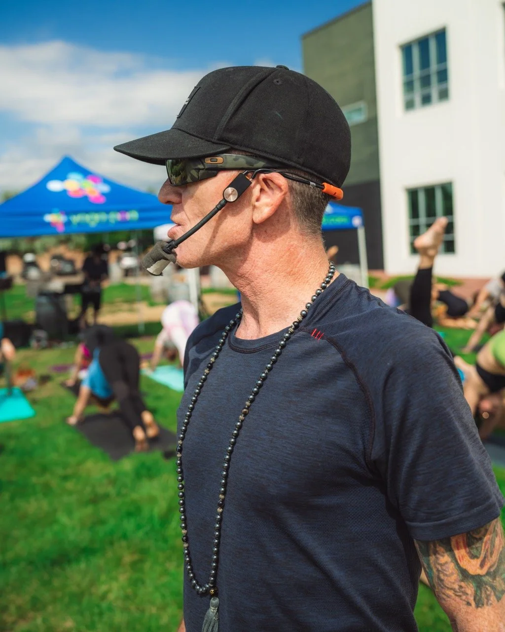 Rob Loud black cap, sunglasses, a headset microphone, and a beaded necklace standing outdoors during a yoga class with people in yoga poses on the grass and a blue tent in the background.