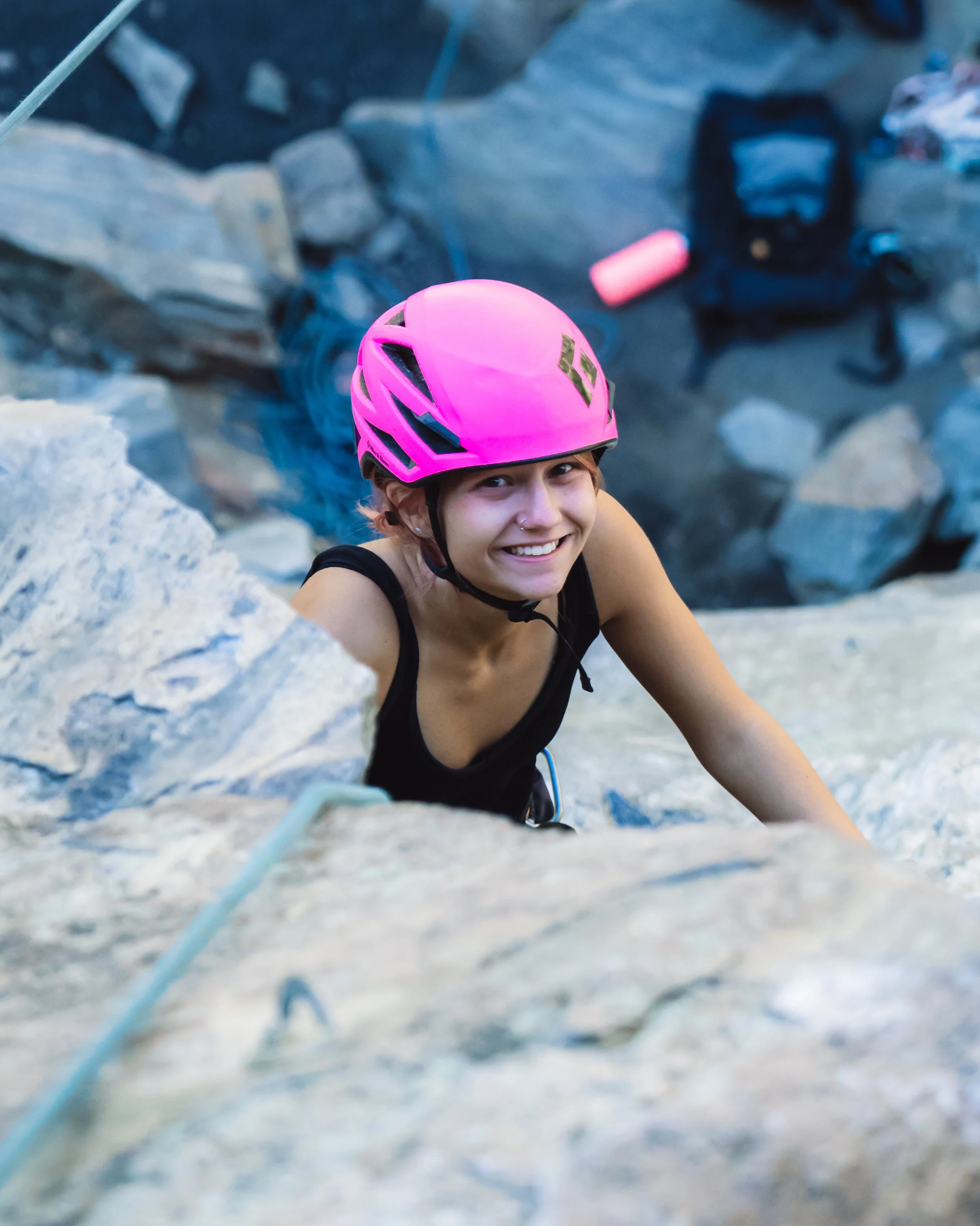 A smiling young woman with a nose piercing climbing a rock wall near Golden, CO wearing a pink helmet and black tank top.