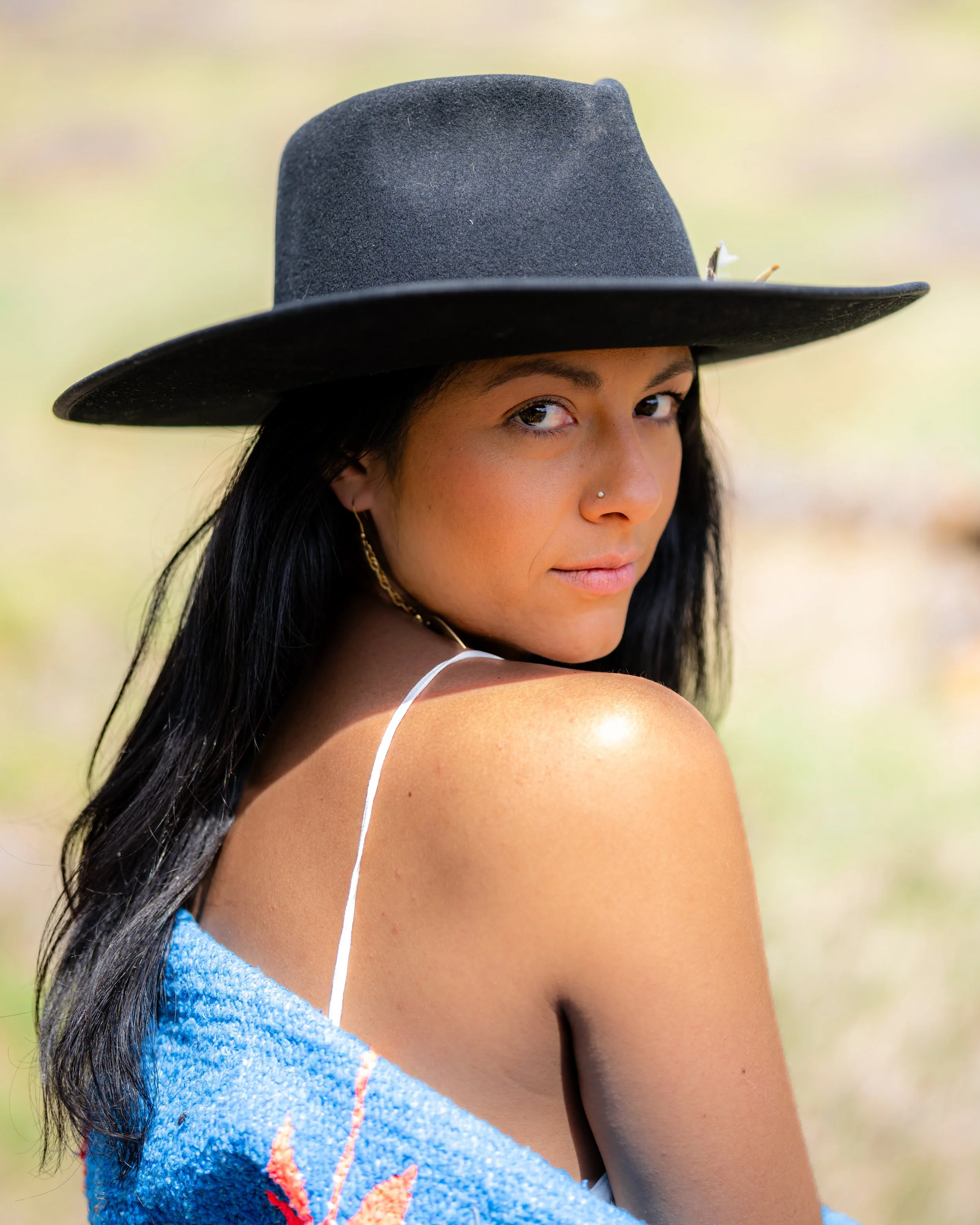 A woman with long black hair wearing a black wide-brimmed hat, a thin white strap top, and a blue blanket draped over her shoulders, looking over her shoulder at the camera with a slight smile.