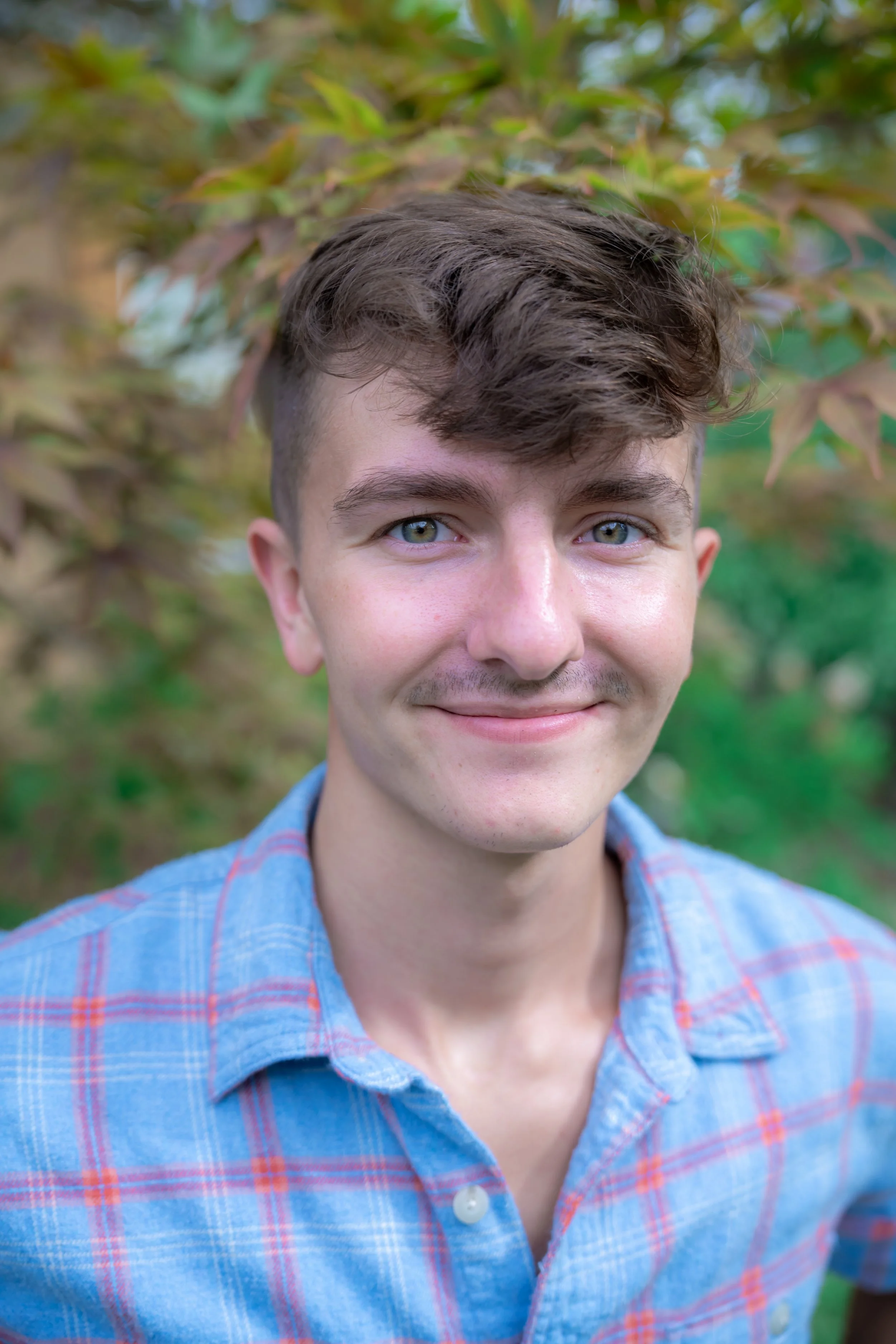 Close-up of a young man smiling outdoors with green foliage in the background.