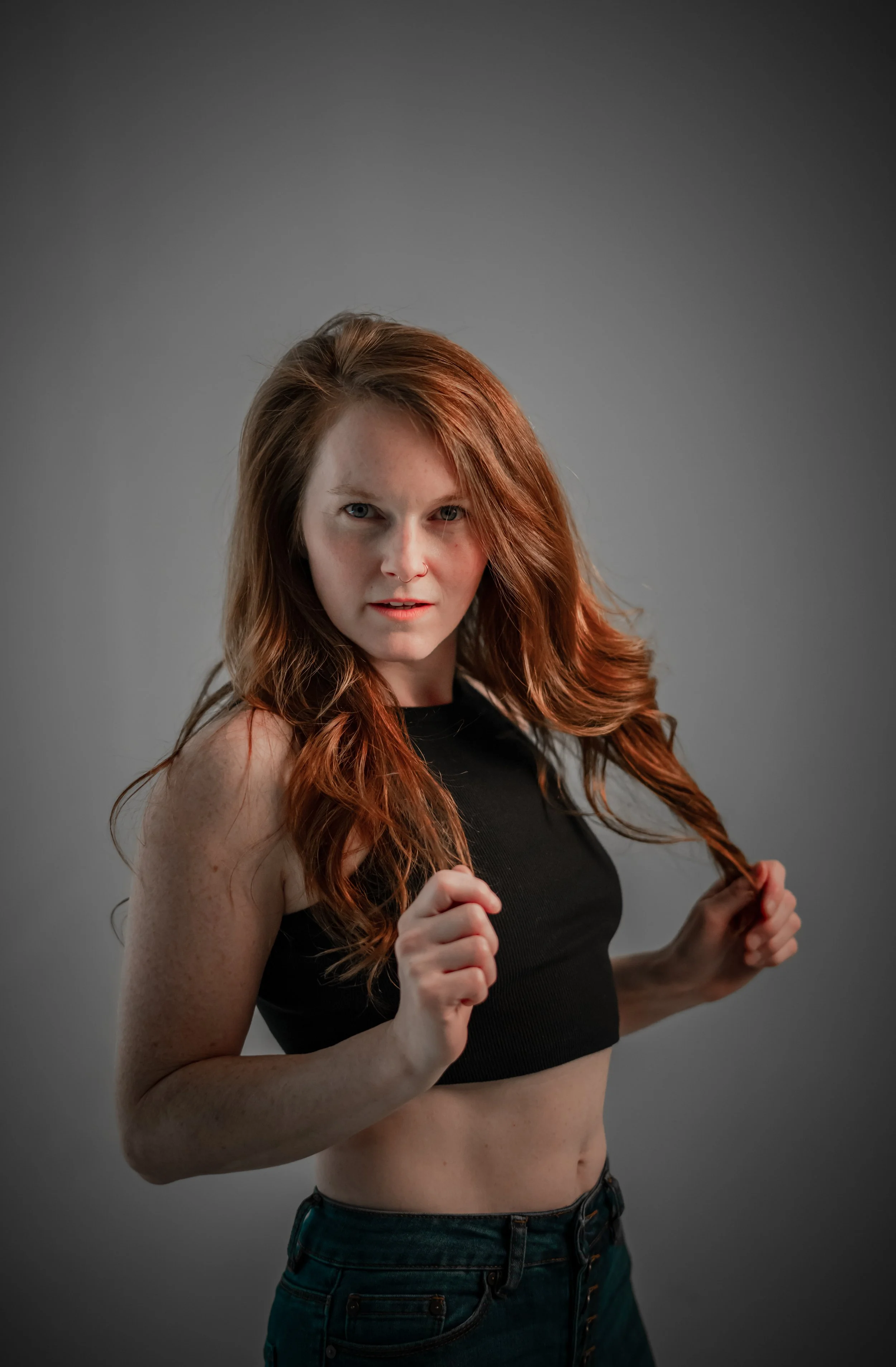 A young woman with red hair, wearing a black crop top and jeans, looking confidently at the camera, holding her hair in both hands against a gray background.