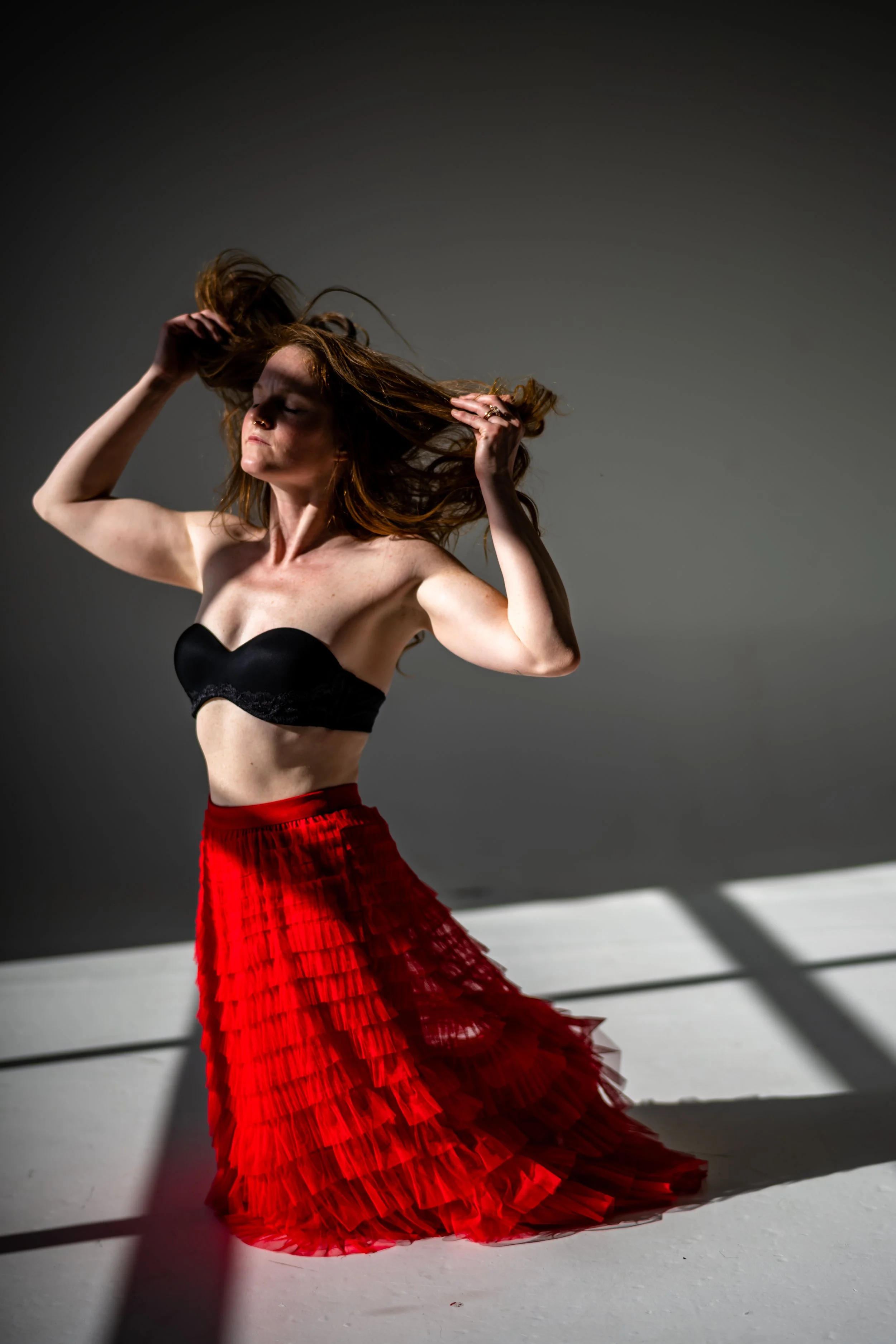 A woman with long red hair wearing a black strapless top and a red ruffled skirt poses with her hands in her hair against a dark background, with natural light casting shadows on the white floor.