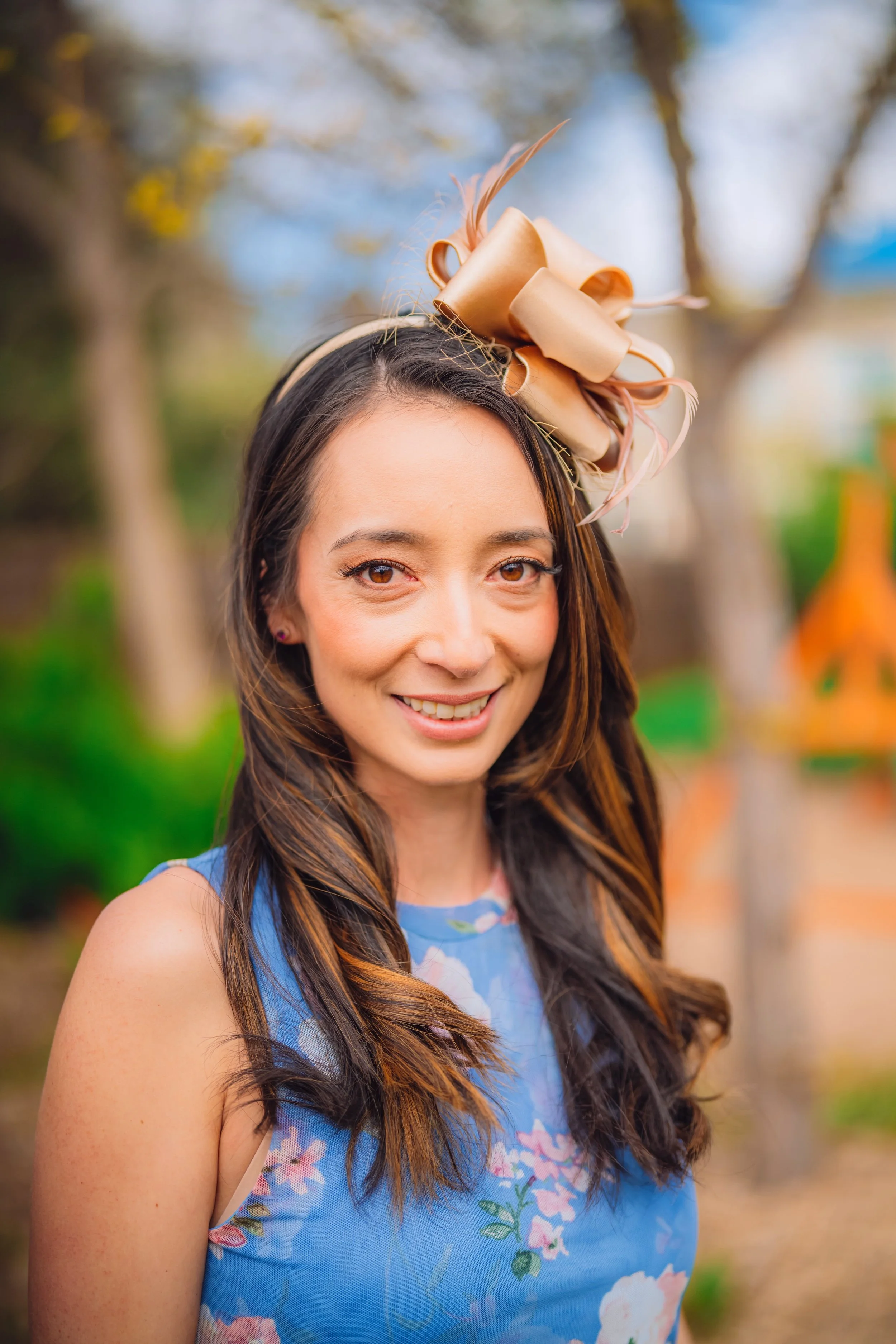 A woman with long brown hair wearing a light blue floral dress and a beige decorative hat with bows and feathers, outdoors with trees.