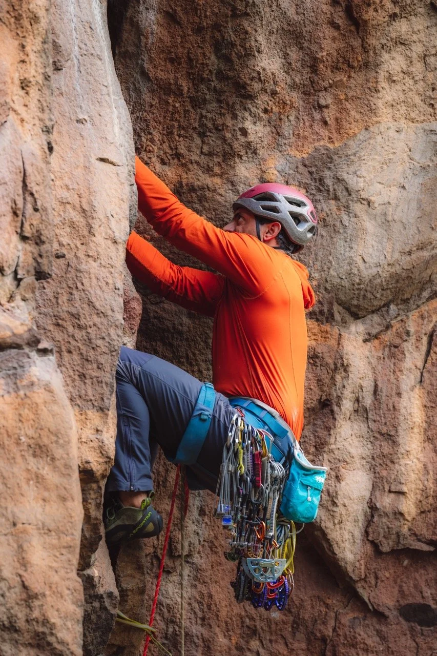 A rock climber wearing an orange jacket and a pink helmet climbing a rock wall, with climbing gear attached to their harness.