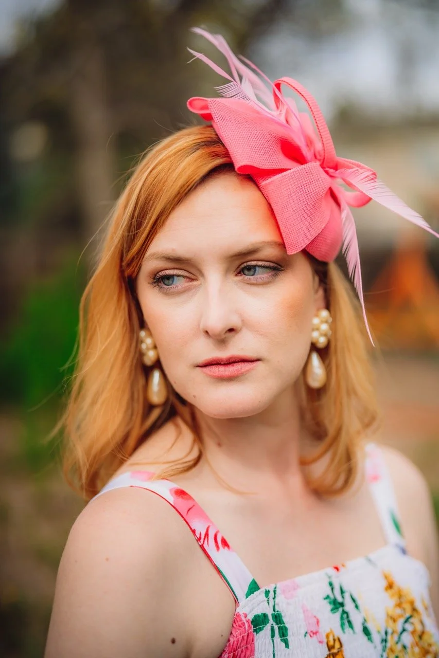 A woman with red hair wearing a large pink fascinator hat, pearl earrings, and a floral dress.
