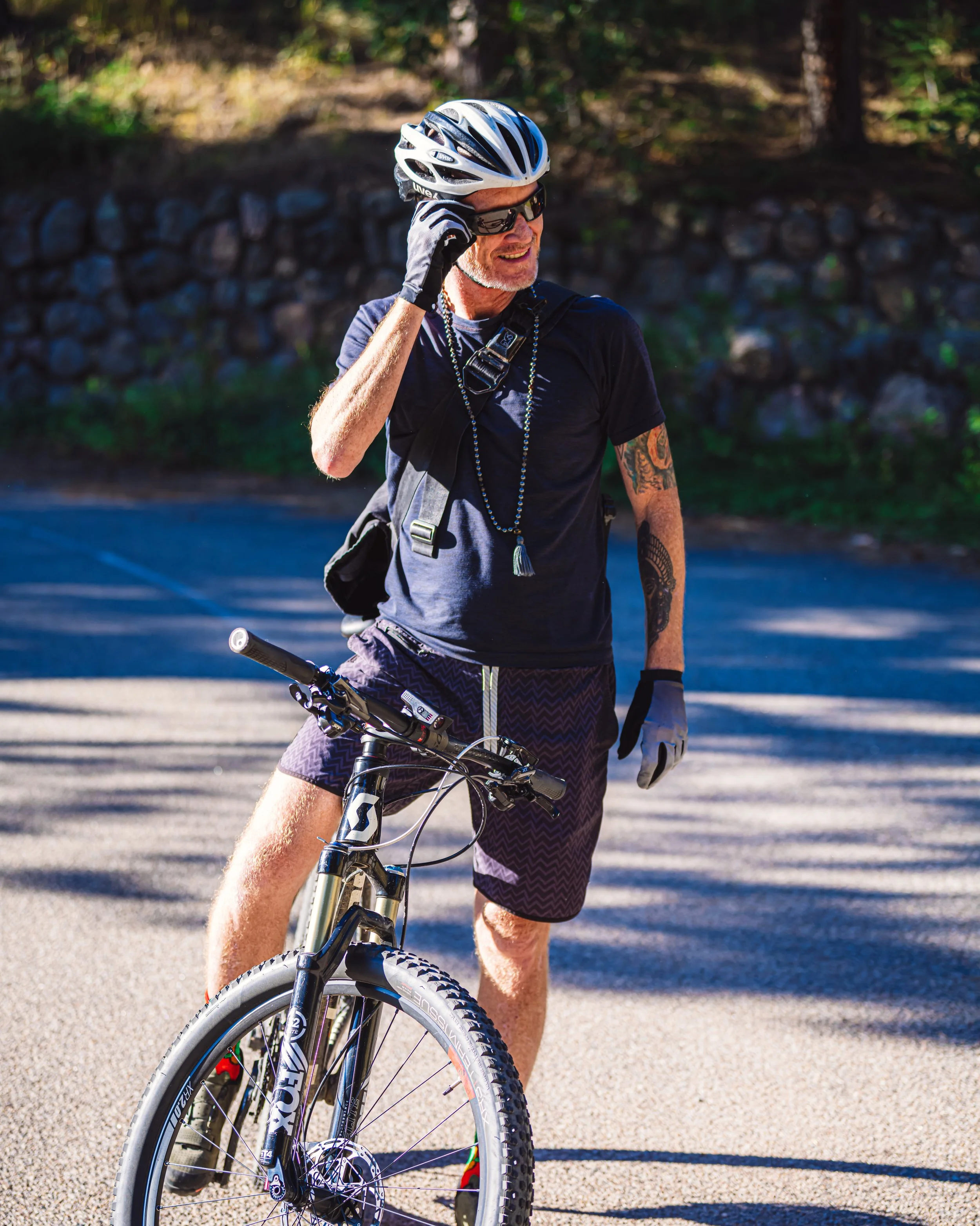Rob Loud in cycling gear, helmet, sunglasses, and gloves standing next to a mountain bike in an outdoor setting with trees and a stone wall in the background.