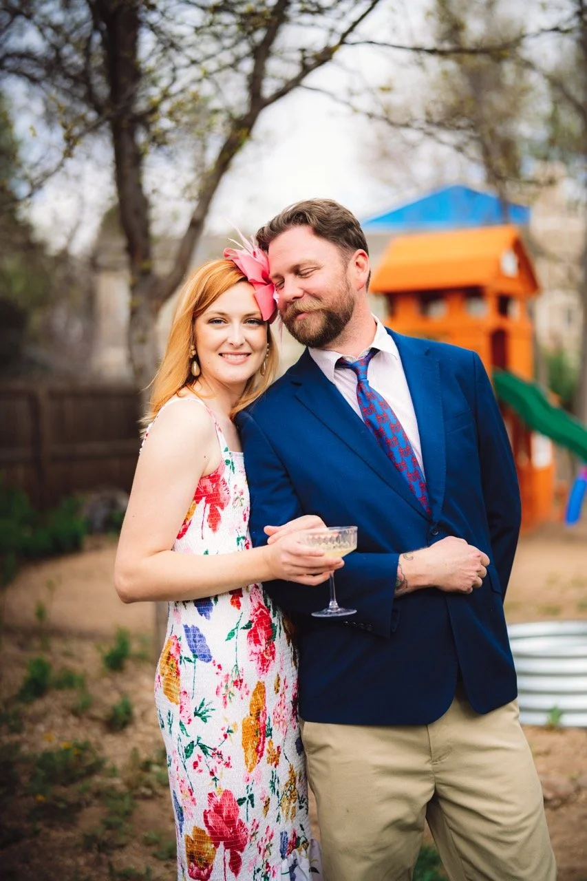 A smiling woman with red hair and a pink bow in her hair, wearing a colorful floral dress, stands next to a man with a beard in a blue blazer, who is holding a drink, in a backyard with a playset in the background.