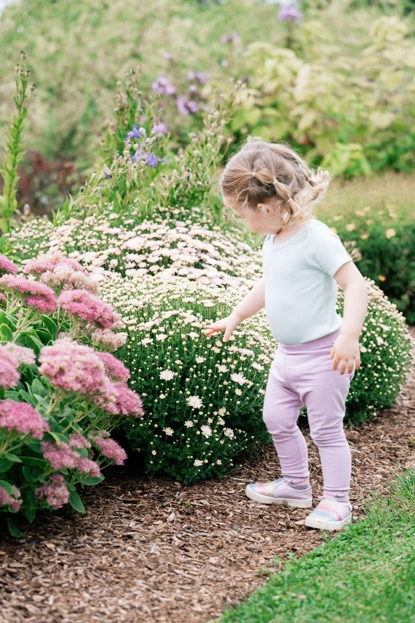 A young girl with blonde hair in a braid, wearing a light blue shirt, pink pants, and pastel sneakers, touching white flowers in a garden with various flowering plants and greenery.