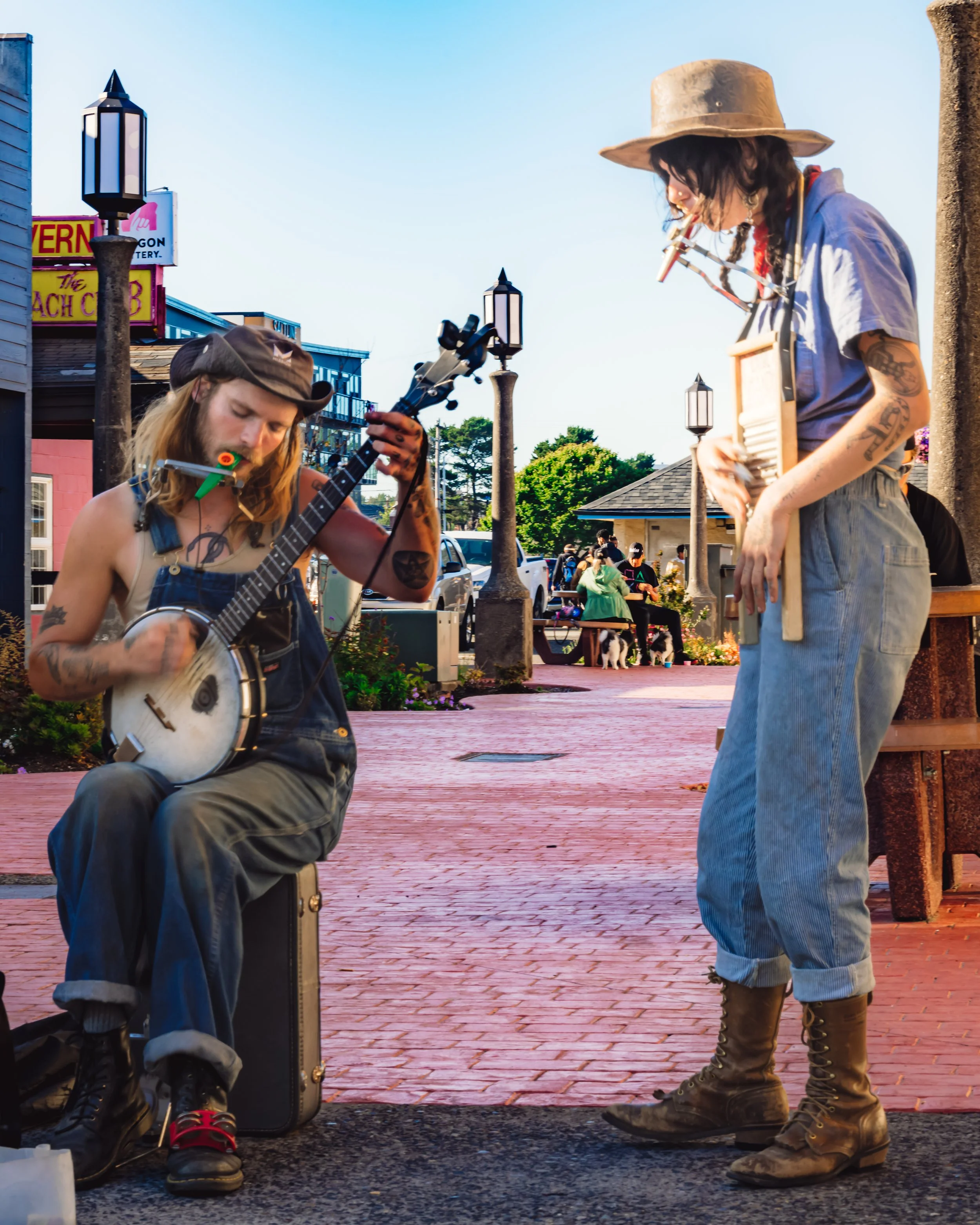 Two street musicians performing outdoors, one playing banjo and the other playing washboard, on a brick sidewalk with lamp posts and people in the background.