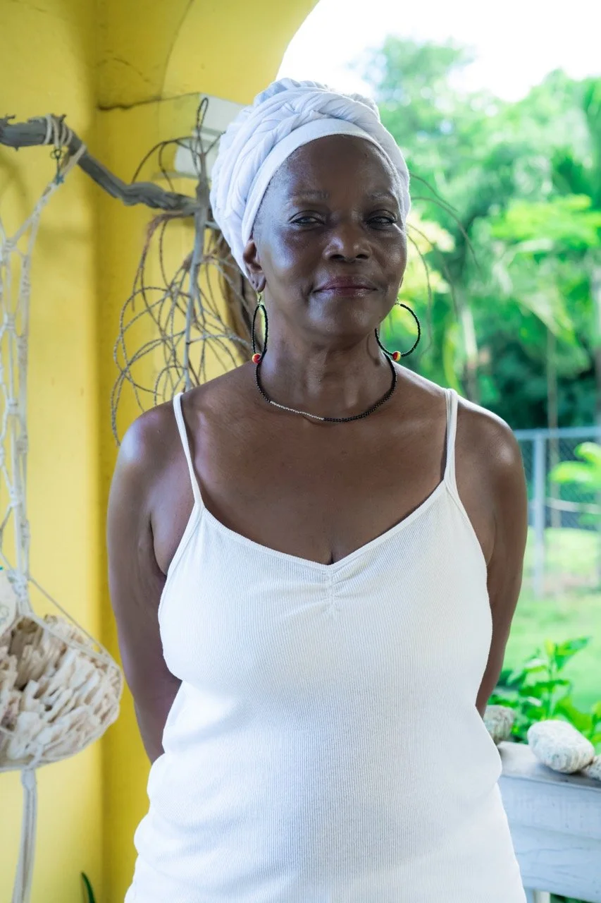 A woman with dark skin wearing a white tank top, black beaded necklace, large hoop earrings, and a white headwrap, standing outdoors near a yellow wall with green foliage in the background.