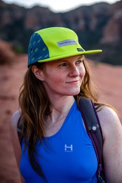 Young woman with long red hair wearing a blue tank top and a yellow and blue cap, outdoors with rocky landscape in the background.