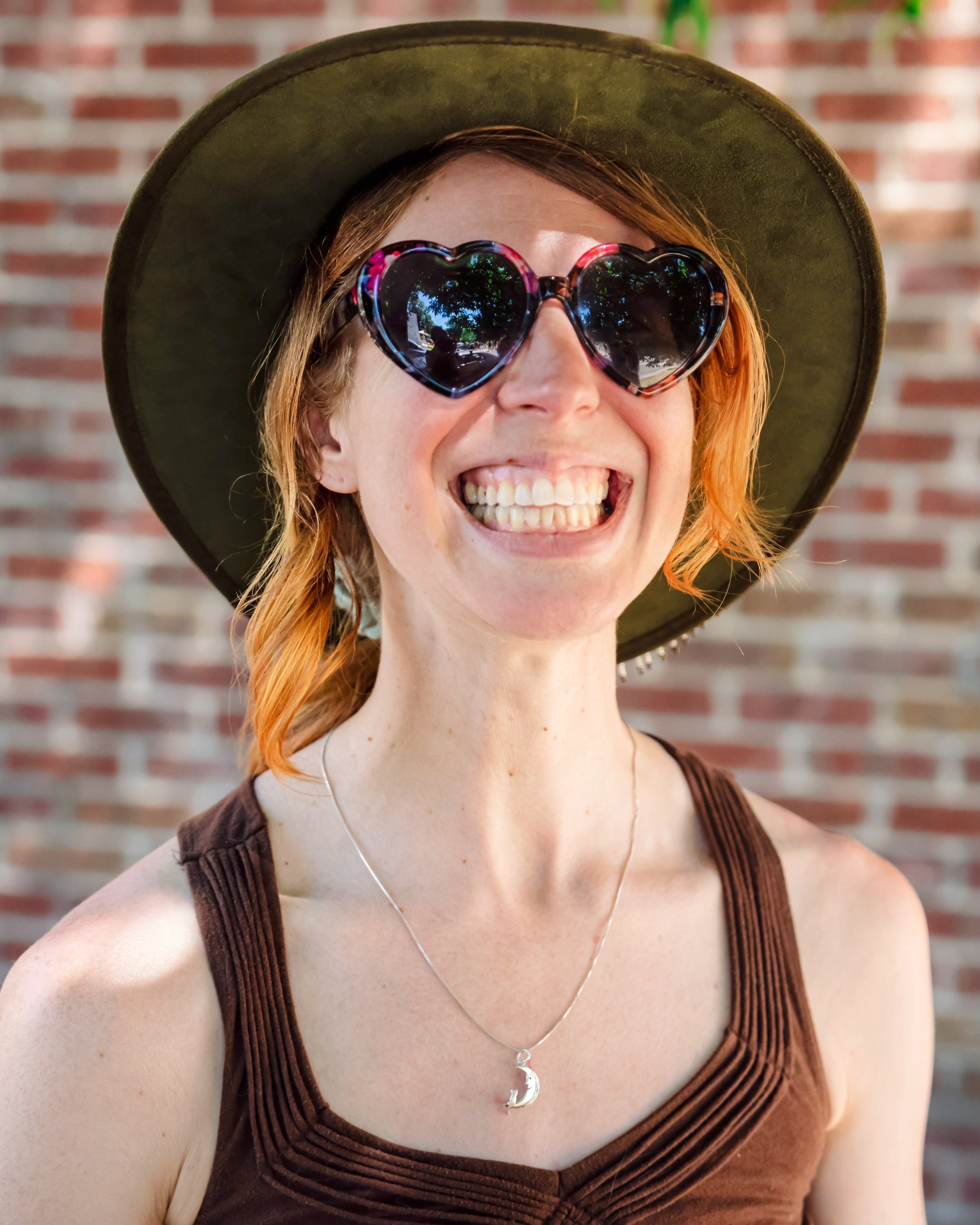A smiling woman wearing heart-shaped sunglasses, a wide-brimmed hat, a brown tank top, and a moon pendant necklace. She has red hair and is standing in front of a brick wall.