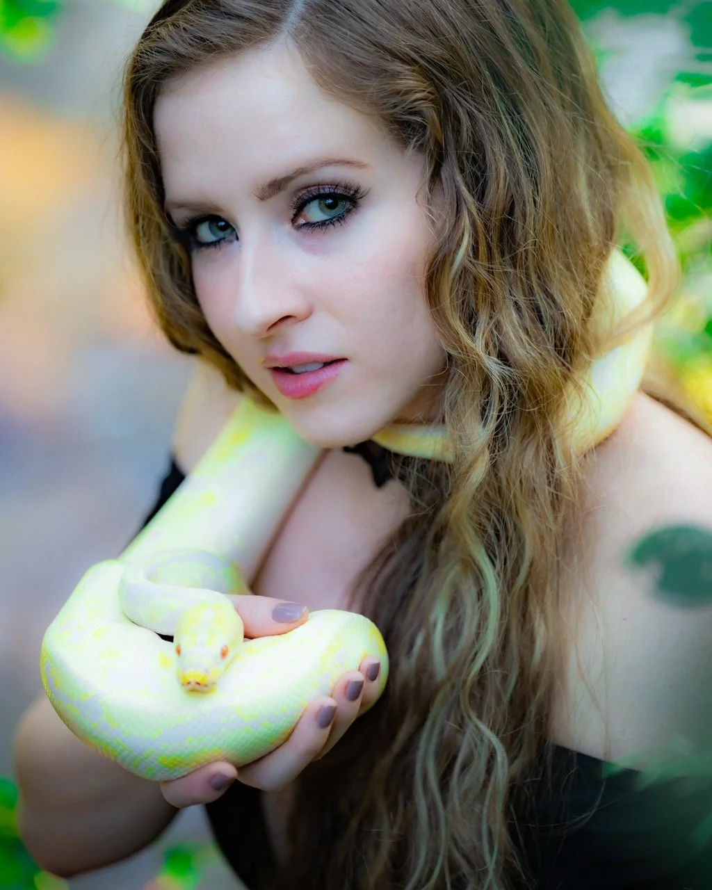 A woman with wavy, long light brown hair and blue eyes holding a yellow and white snake around her shoulders and hand, outdoors with green foliage in the background.