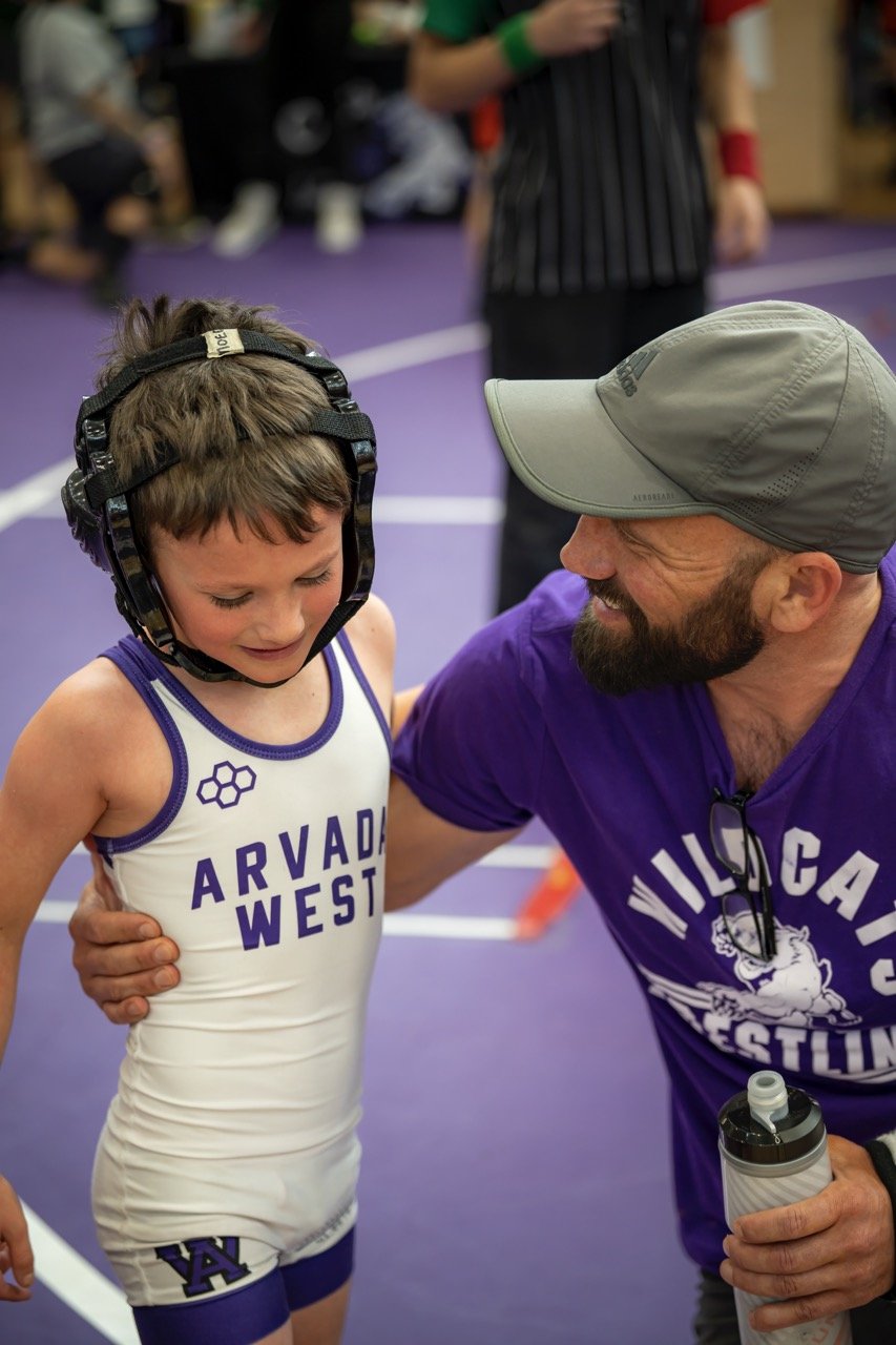 A young male wrestler in a white and purple Arvada West singlet wearing a black protective headgear is talking to an adult male coach or trainer in a purple shirt and gray cap, inside a wrestling gym with purple mats.