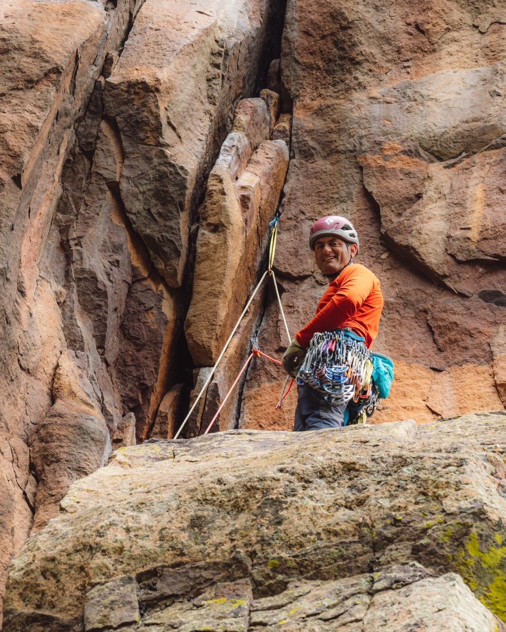 A male rock climber wearing a helmet and climbing gear standing on a rocky ledge in Eldorado Park, CO, smiling at the camera, with a climbing rope attached to his harness in front of a rugged rock face.