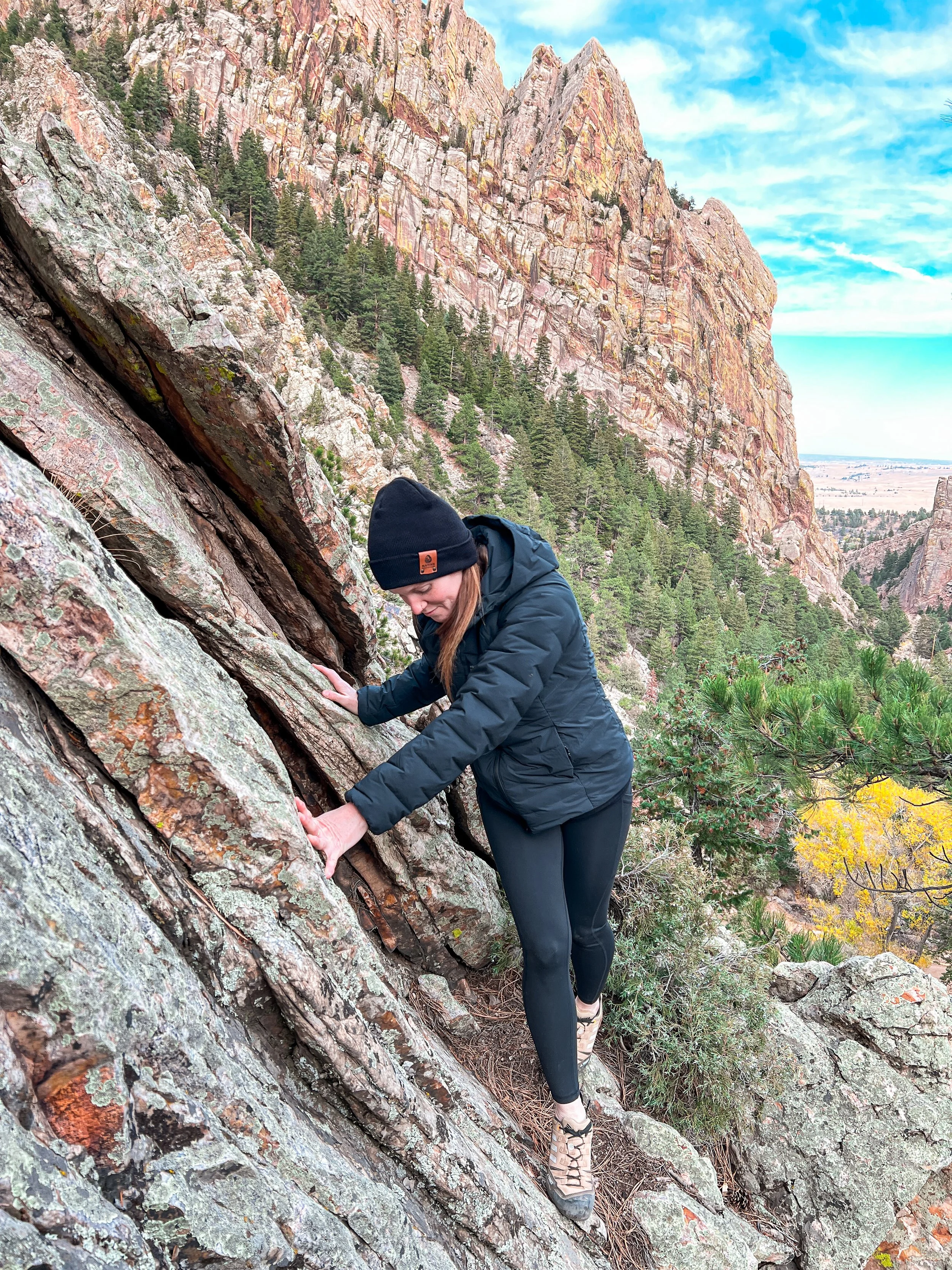 Person wearing a black beanie and dark jacket climbing a steep rock face in a mountainous area with pine trees and colorful rock formations in the background.