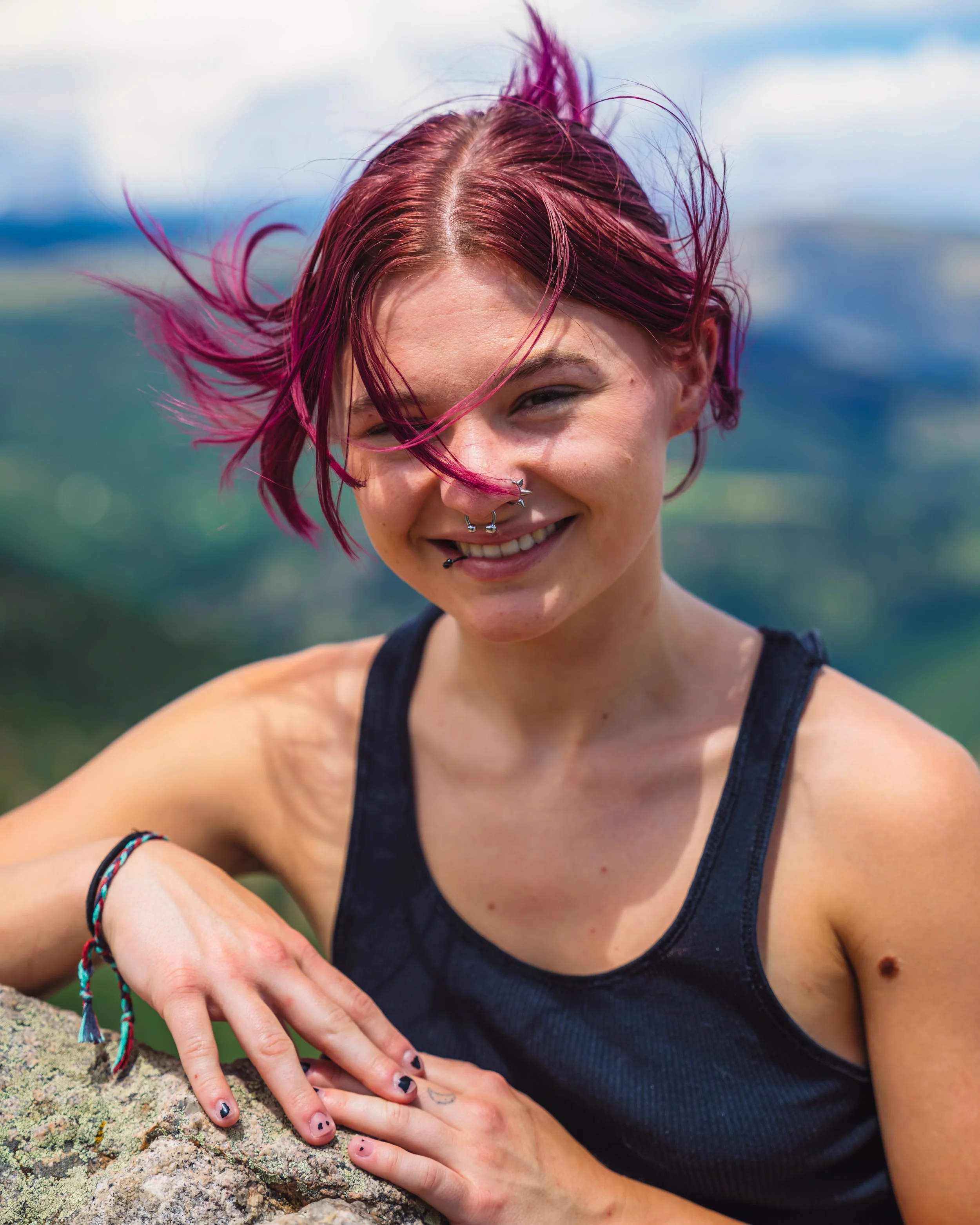 A young woman with pink and purple hair, wearing a black tank top, smiling outdoors with mountains and greenery in the background.