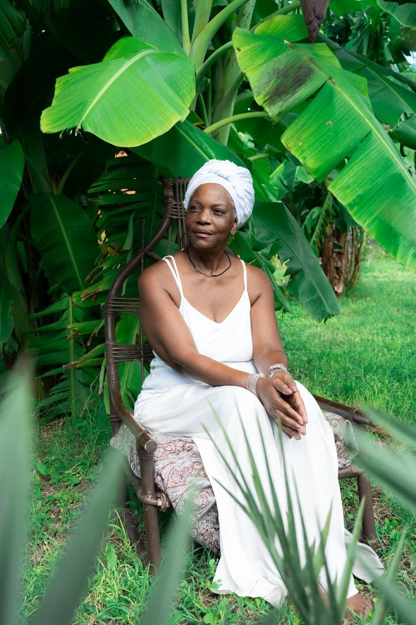 A woman dressed in white sitting on a wooden chair in a lush green garden with large banana leaves surrounding her.