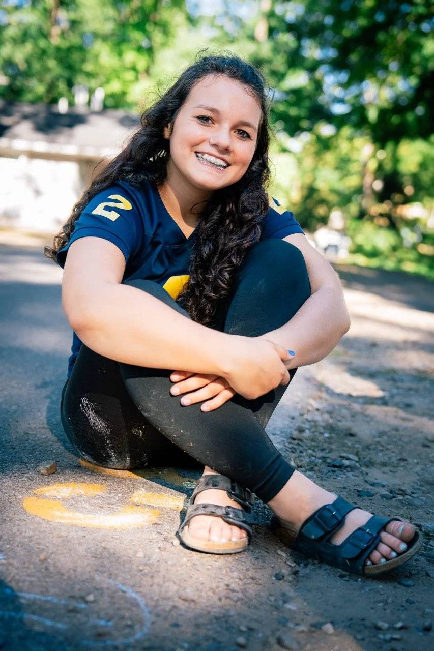 A smiling girl with long curly hair and braces, sitting outdoors on the ground, wearing a blue sports jersey, black leggings, and sandals, with trees and a sunny sky in the background.