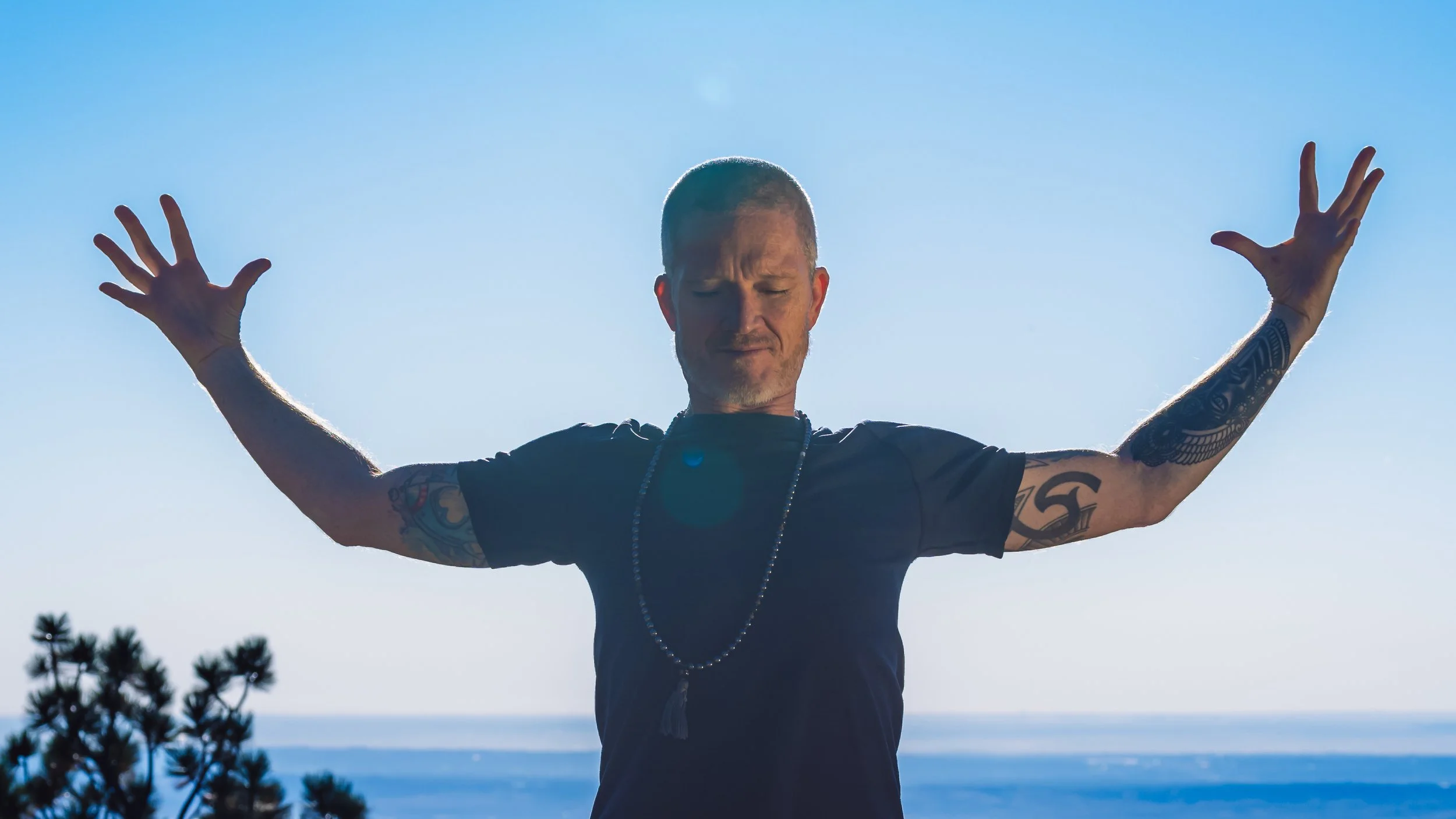 Rob Loud stands before the city of Boulder with arms raised and eyes closed, with a clear blue sky and distant ocean in the background.