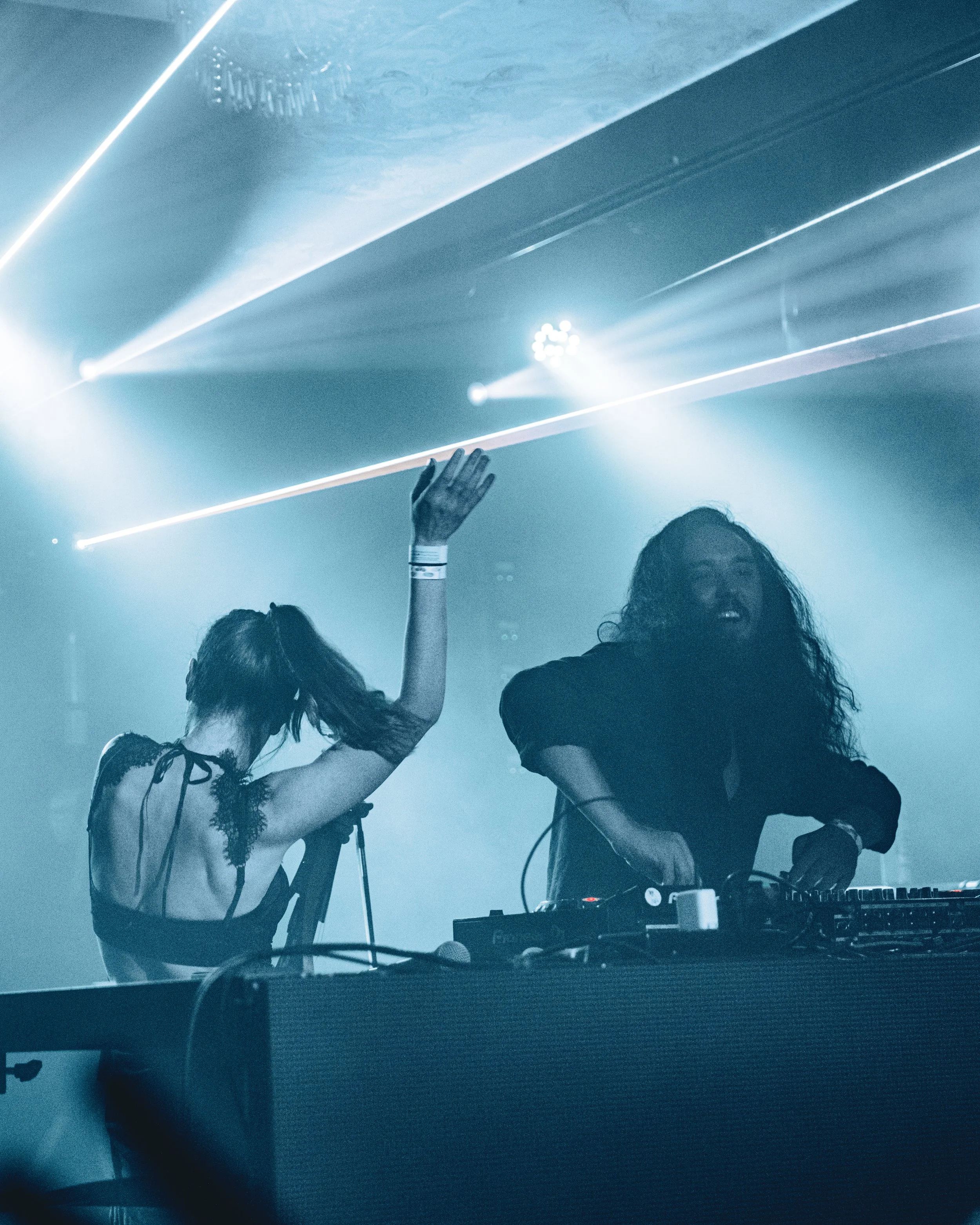 Denver duo Spectacle performs at Cervantes' Other Side with laser lights and fog, Michael turning knobs on the equipment and Jessica with her back to the camera, raising her hand in dance.
