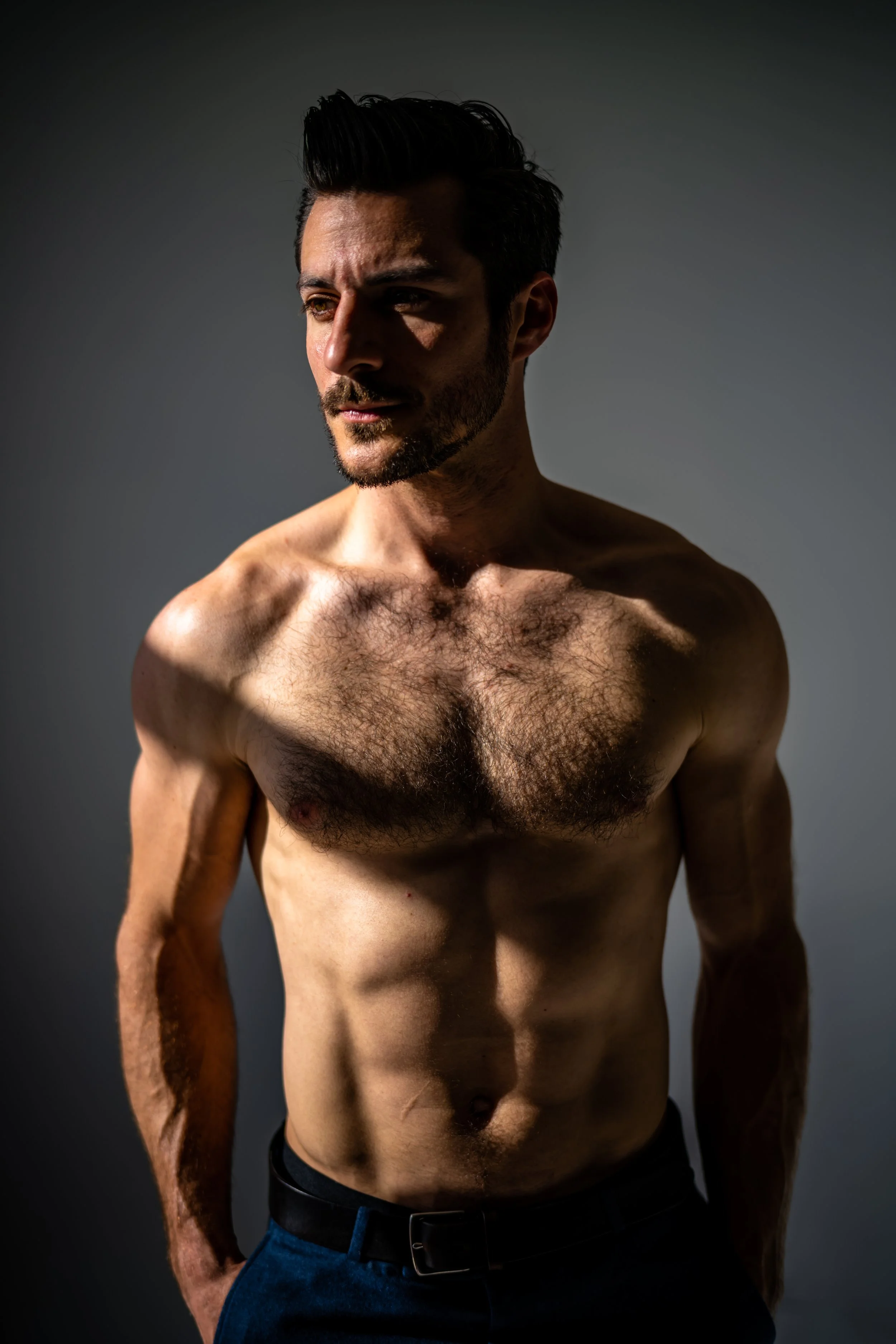 A shirtless man with dark hair and beard stands against a plain gray background, looking slightly to the side with shadowed natural light highlighting his muscular physique and chest hair.