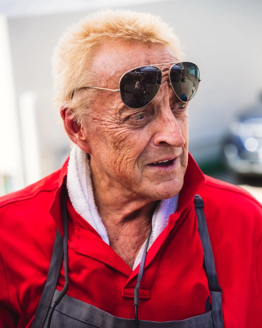 An elderly man with fair skin, blonde hair, and glasses, wearing a red jacket and a white towel around his neck, standing outdoors.